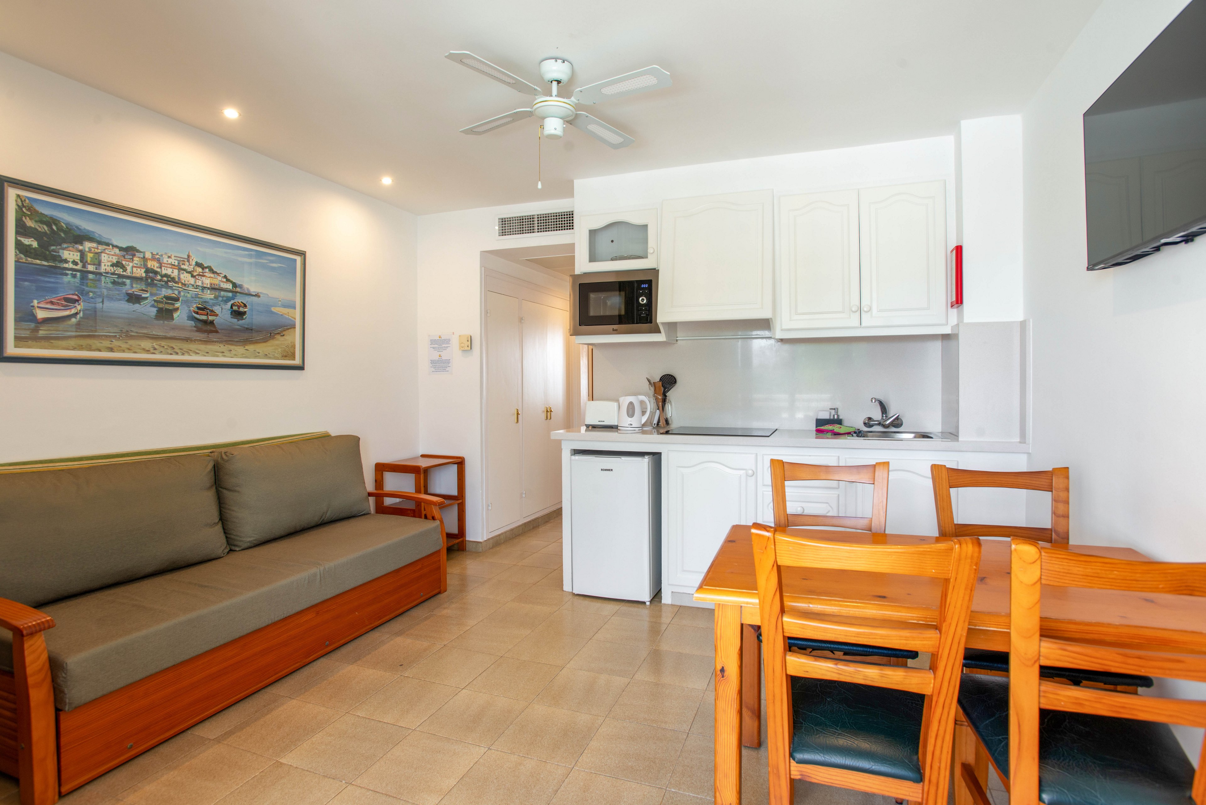 Compact studio apartment interior featuring a small kitchenette and dining area. A wooden dining table with chairs sits beside a white counter with sink, microwave, kettle, and mini fridge. A simple wooden sofa with gray cushions lines the opposite wall under a coastal landscape painting. Neutral walls, tiled floor, ceiling fan, and recessed lighting create a clean, bright atmosphere.