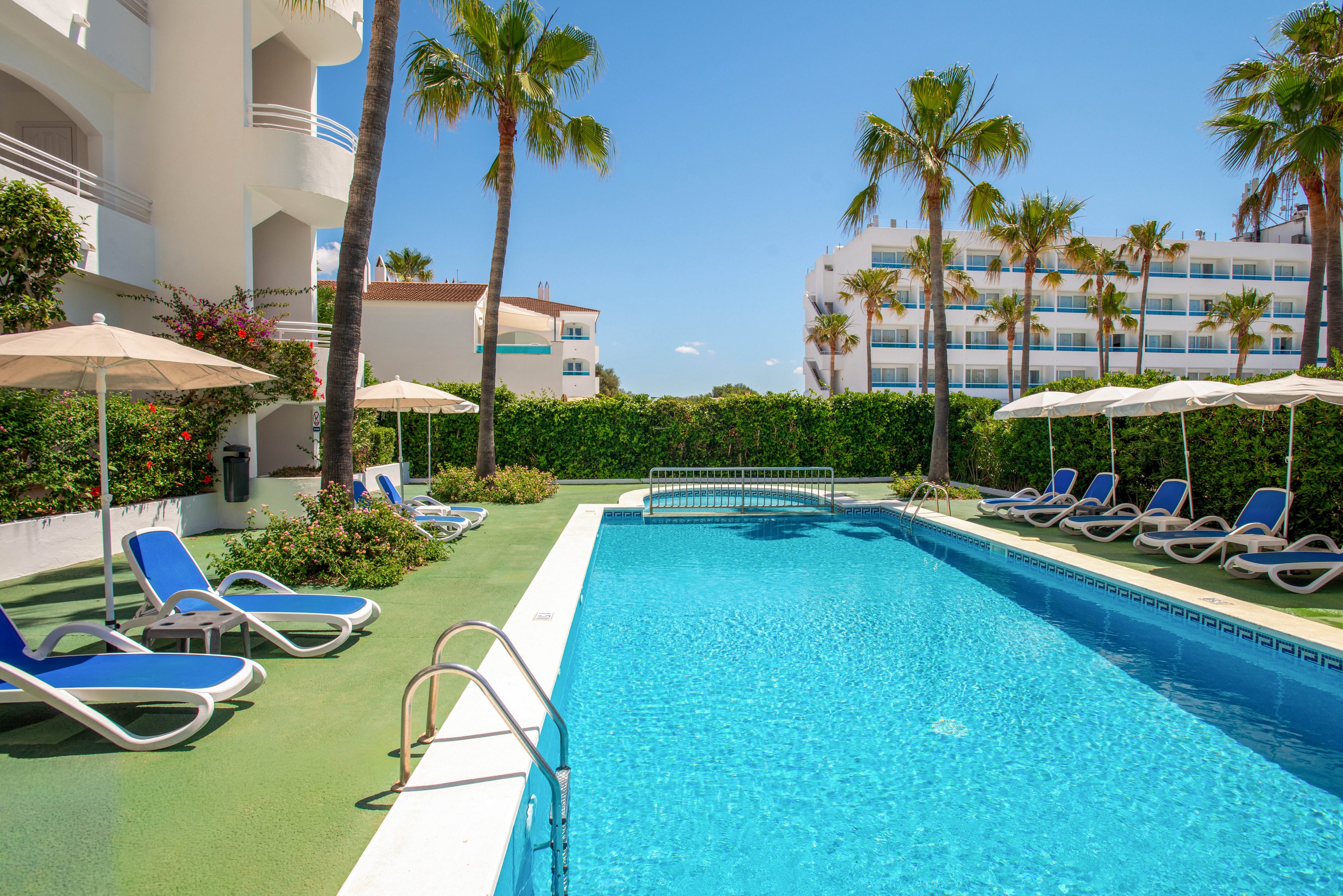 An inviting outdoor swimming pool is surrounded by blue sun loungers and beige umbrellas at a modern hotel. Tall palm trees and manicured hedges frame the pool area, creating a resort-like atmosphere. White multi-story buildings with balconies rise in the background under a clear blue sky. The bright sunlight and vivid colors suggest a warm, relaxing vacation setting.
