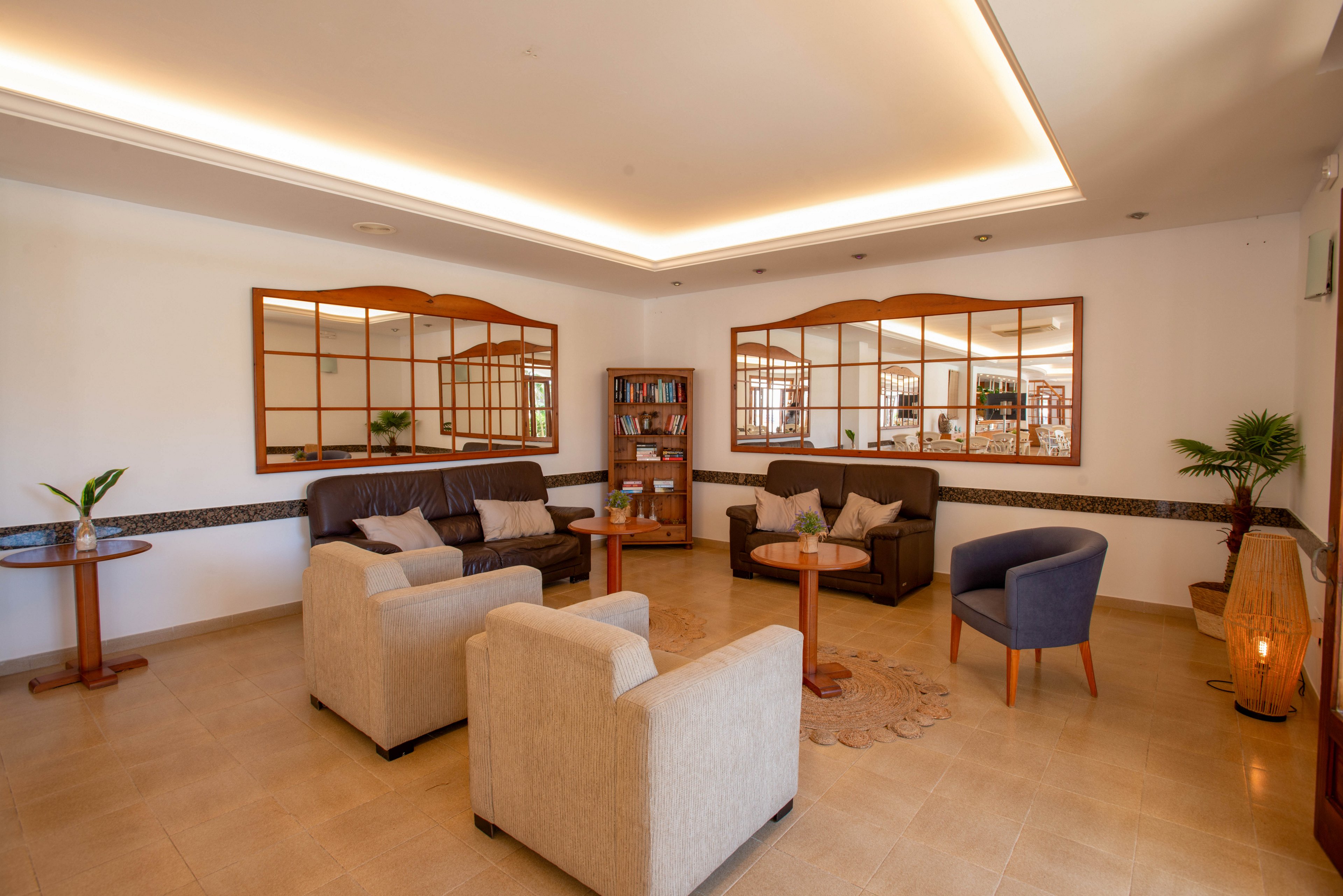 A warm, inviting lounge area features several armchairs and sofas arranged around small wooden tables. Large wall mirrors reflect the room, adding depth and light to the neutral-toned space. Potted plants, bookshelves, and soft lighting create a relaxed, comfortable atmosphere. The clean tiled floor and minimalist decor suggest a modern hotel or guesthouse interior.