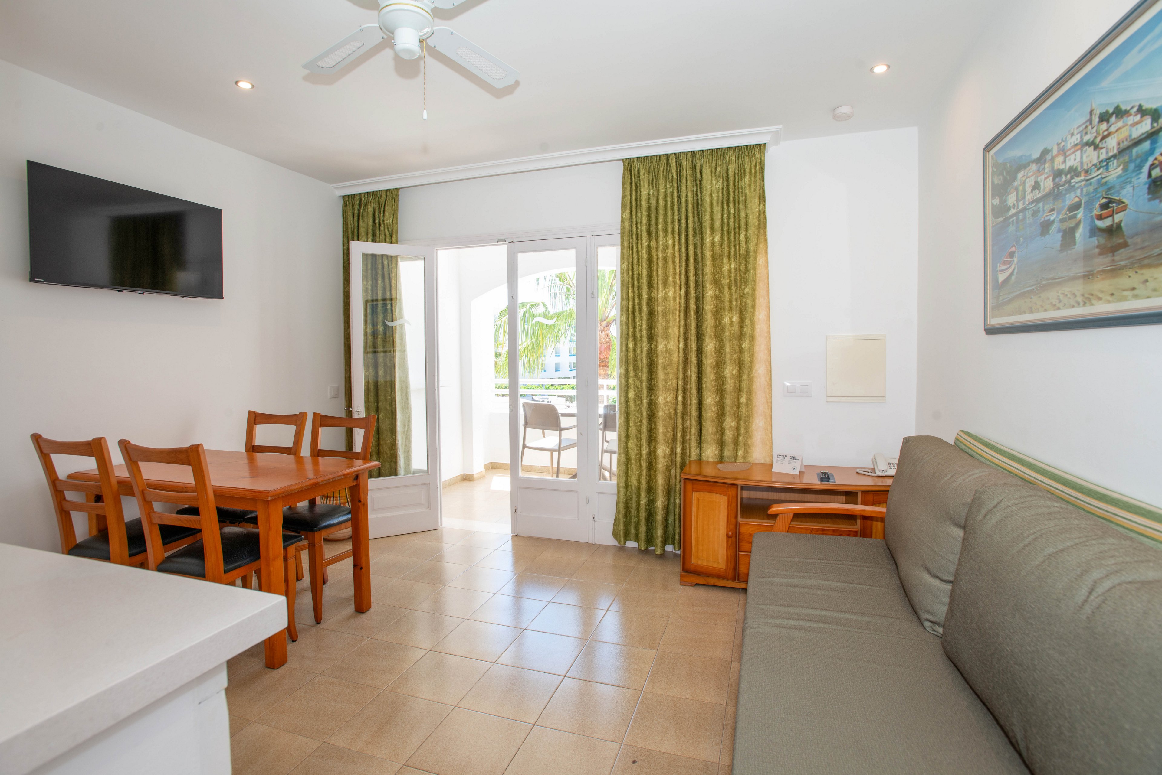 A clean, modern apartment living room features a gray sofa and a wooden dining table with four chairs. Large glass doors open onto a sunny balcony, allowing natural light to fill the tiled space. A flat-screen television is mounted on the wall opposite the seating area, and green curtains frame the doorway. A ceiling fan and coastal artwork contribute to the relaxed vacation atmosphere.