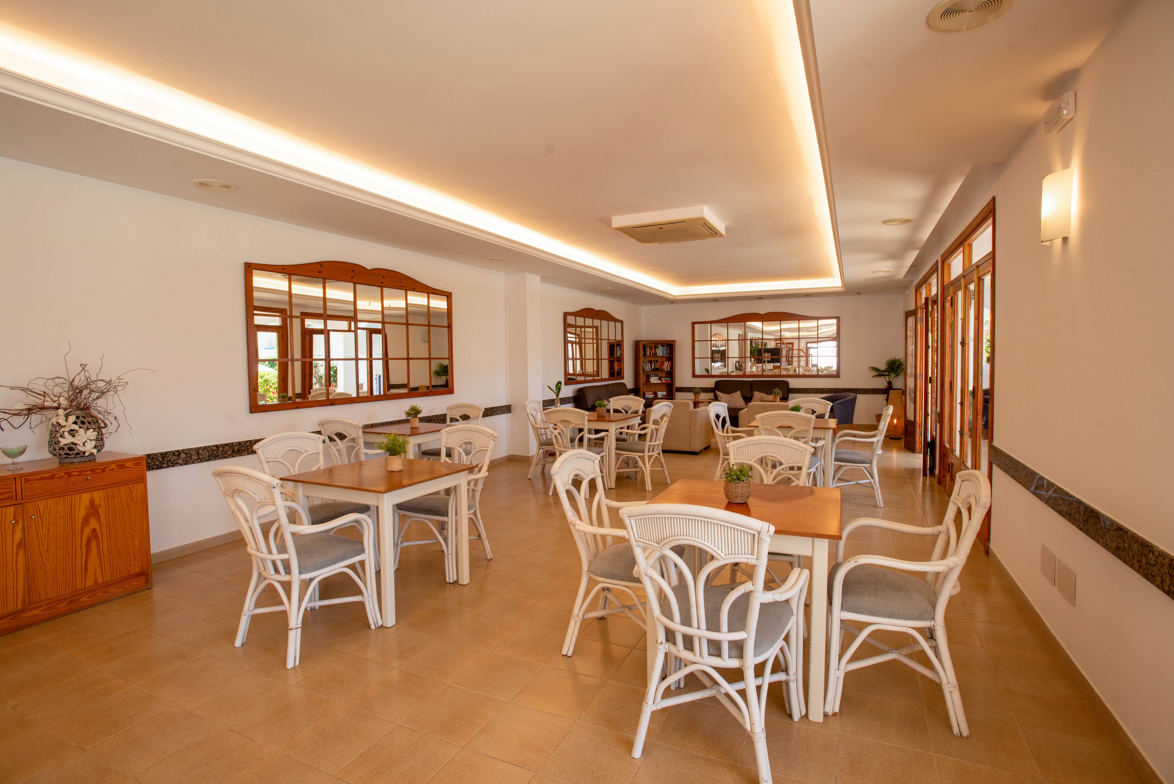 Spacious indoor dining area with multiple tables and white wicker chairs arranged neatly. Warm ceiling lighting and large wooden-framed windows create an inviting atmosphere. A sideboard with decorative plants and a wall of mirrors add visual interest to the room. The space appears clean, quiet, and ready to welcome guests.