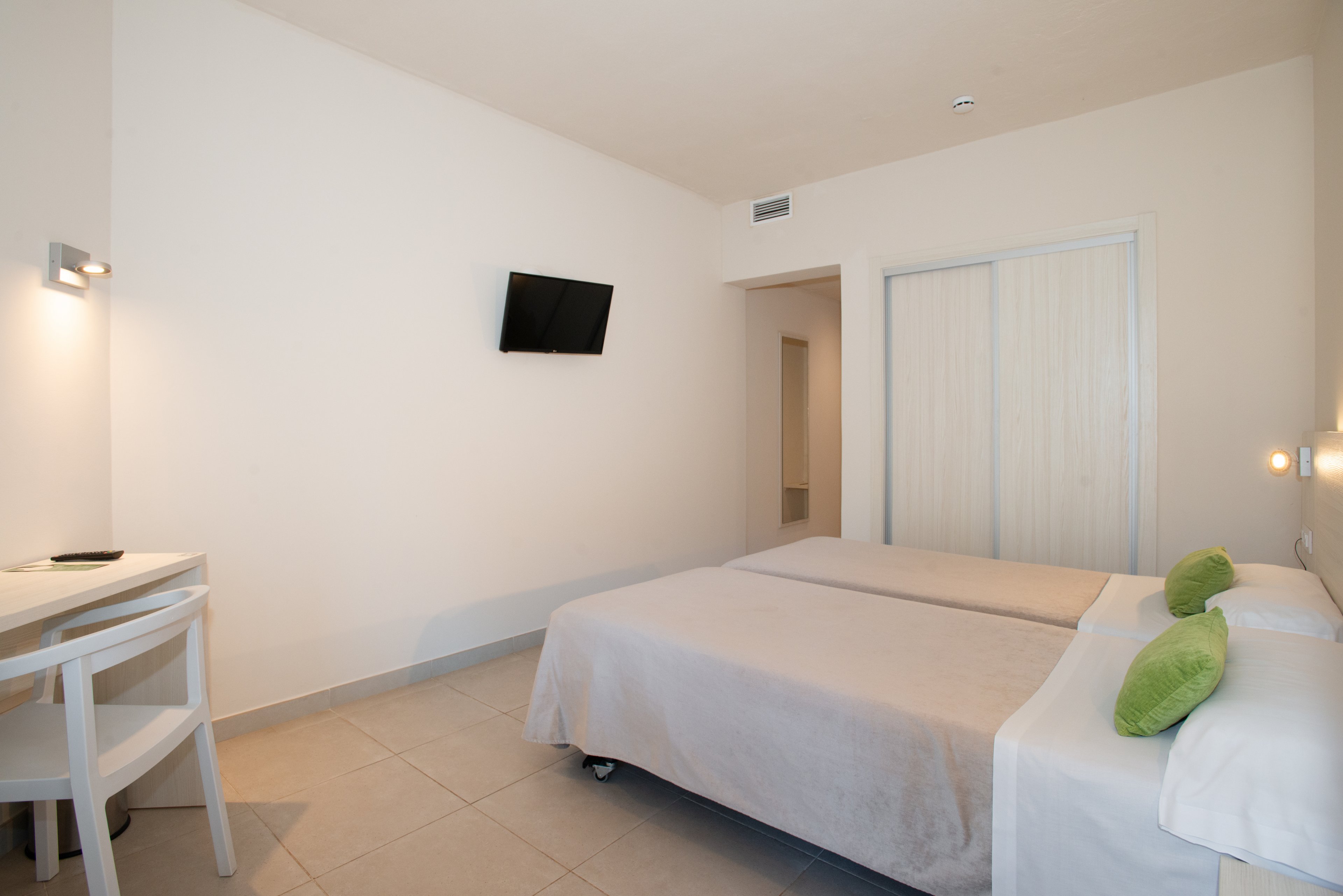 Bright, modern hotel room featuring two neatly made twin beds with light beige bedding and green accent pillows. A small white desk and chair sit in the corner opposite a wall-mounted flat-screen television. Sliding closet doors and neutral tiled flooring contribute to the clean, minimalist aesthetic. Soft wall lighting and pale walls create an airy, uncluttered atmosphere suitable for guests.