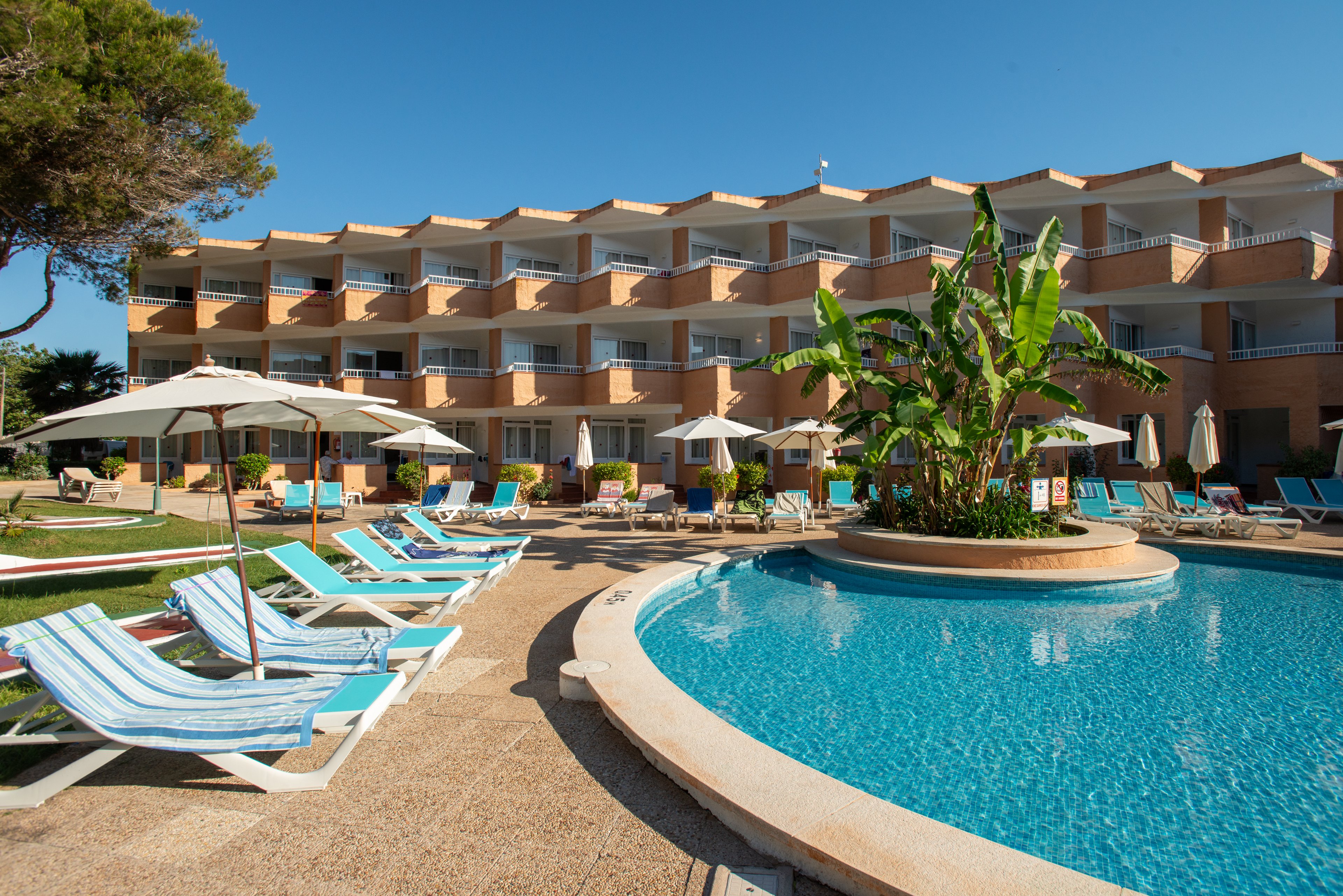 An outdoor swimming pool curves through a sunny resort courtyard lined with blue-and-white lounge chairs. Multiple white sun umbrellas provide shade beside the pool, with green lawn and tropical plants adding color. A multi-story hotel building with repeating balconies forms the backdrop under a clear blue sky. The scene suggests a relaxing vacation atmosphere at a warm-weather destination.