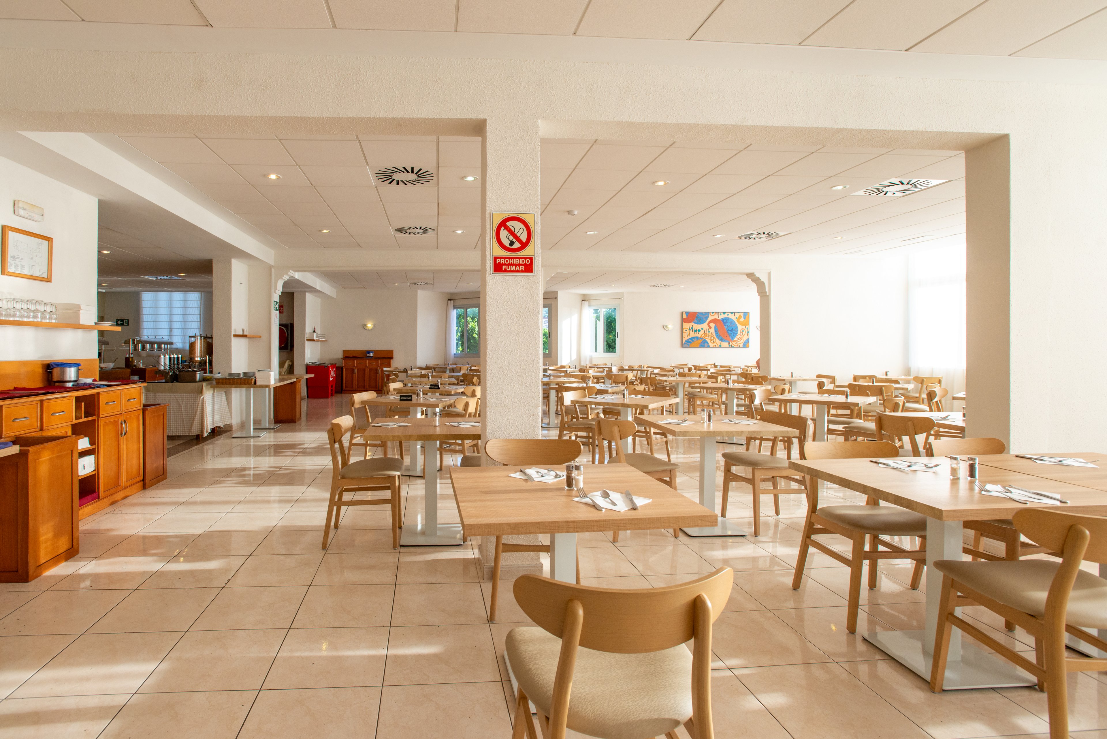 The image shows a spacious, empty cafeteria or dining hall with rows of light wooden tables and chairs. Sunlight streams in through large windows on the right, creating a bright and welcoming atmosphere. A serving area with counters and cabinets is visible on the left side, while a colorful wall painting decorates the far wall. A red circular no-smoking sign is clearly visible on a column near the center of the room.