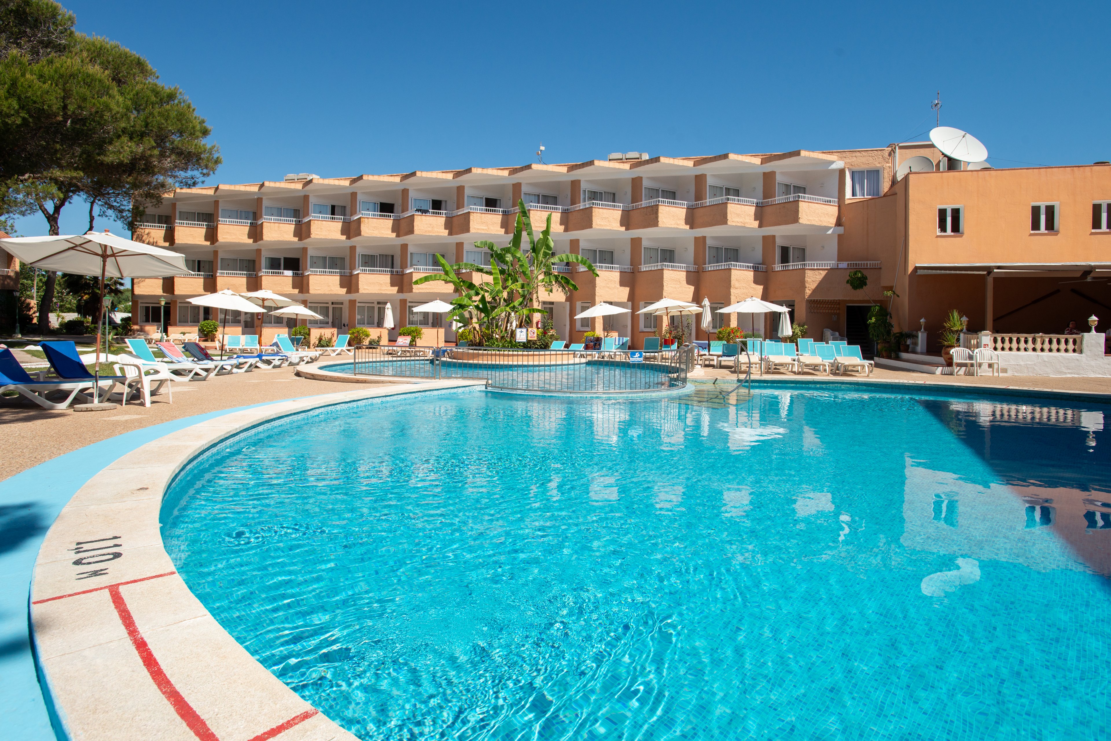 A large outdoor swimming pool is featured in front of a multi-story resort hotel under a clear blue sky. Sun loungers with colorful cushions line the poolside terrace, suggesting a relaxing vacation setting. Tropical plants and trees add greenery around the area, while balconies overlook the pool. The pool edge shows a depth marking of 1.40 m near the foreground.