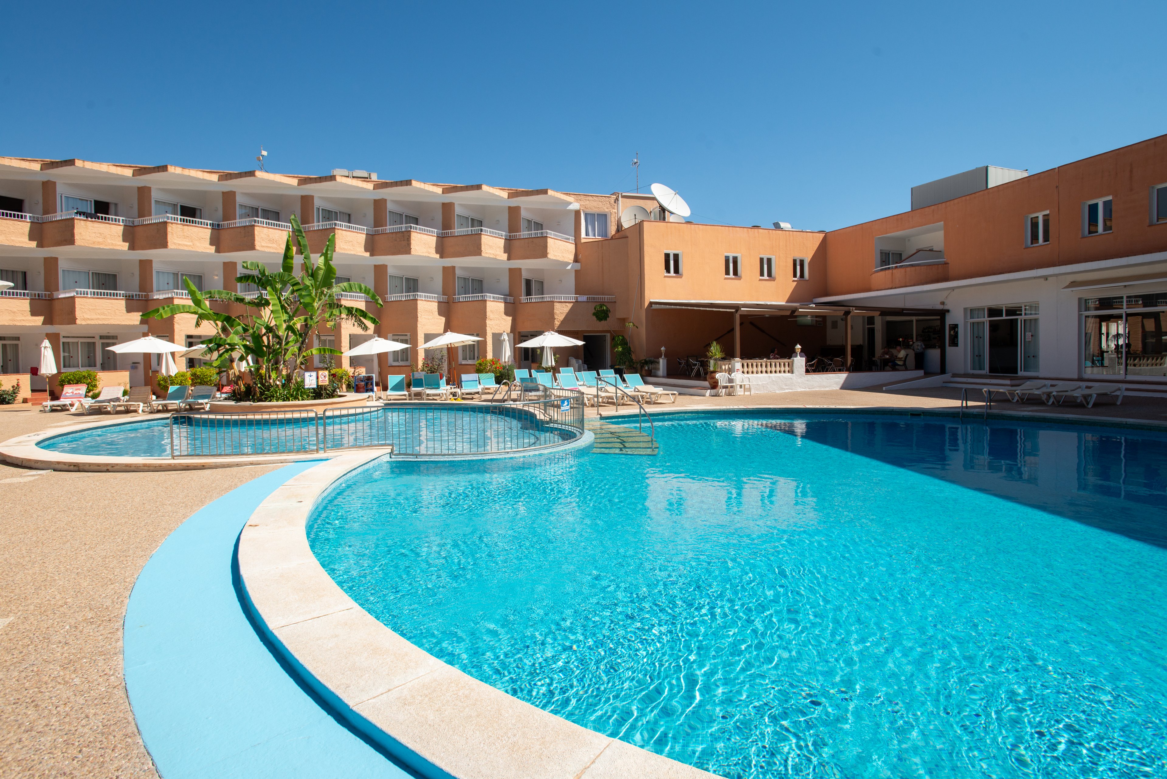 A large outdoor swimming pool sits at the center of a hotel courtyard under a clear blue sky. The multi-story building features balconies overlooking the pool area, with sun loungers and umbrellas arranged along the deck. A small circular pool section is connected to the main pool, and tropical plants add greenery to the scene. The bright sunlight and clean architecture create a relaxed vacation atmosphere.