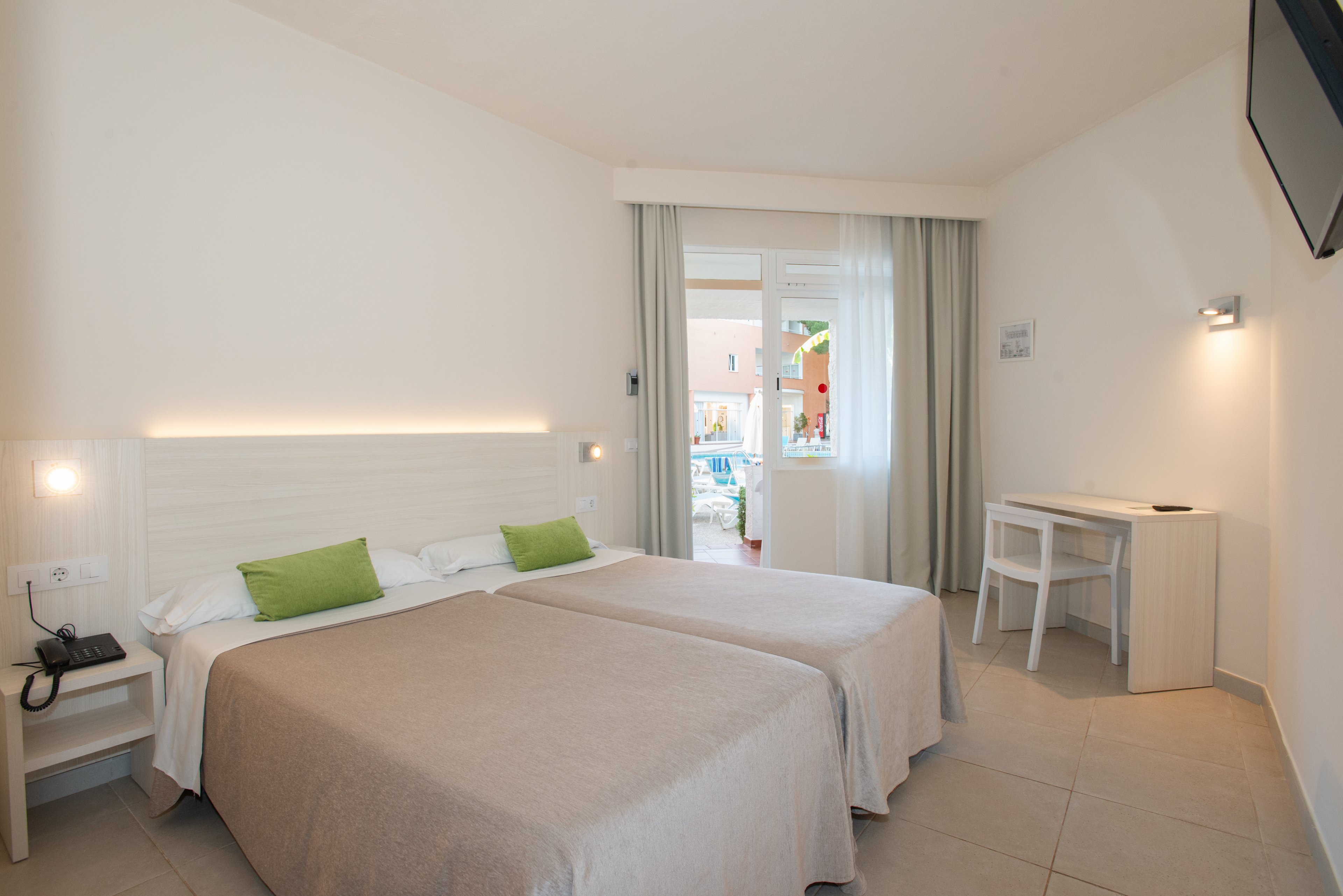 A modern hotel room features two twin beds with beige covers and green accent pillows against a light-colored wall. A small desk and chair sit to the right, beneath a wall-mounted television. Natural light enters through glass doors leading to a balcony with a view of an outdoor pool area and buildings. The room has a clean, minimalist design with neutral tones and simple decor.