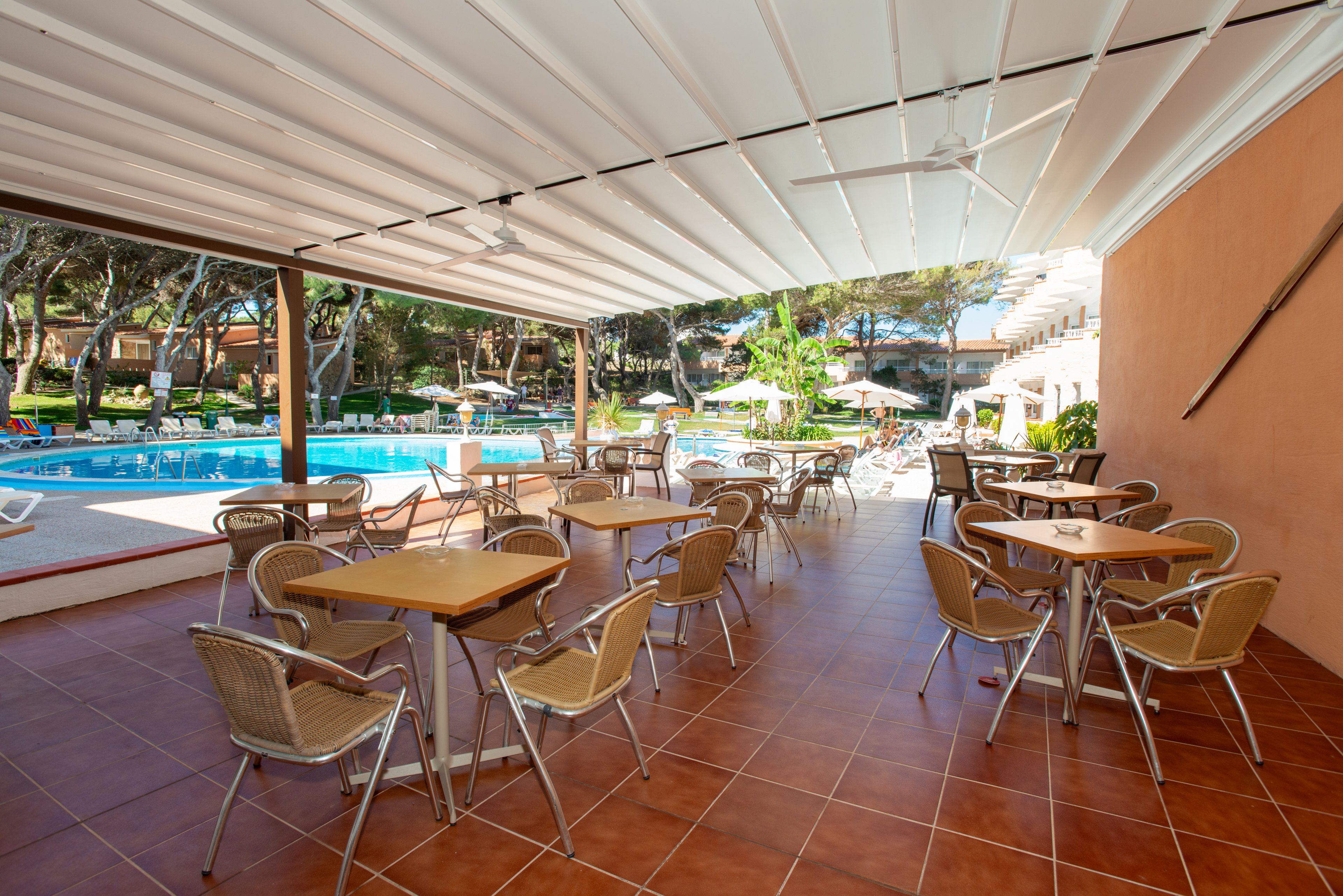 An open-air café terrace with metal chairs and wooden tables overlooks a bright blue swimming pool. The area is covered by a white canopy roof, providing shade over the tiled floor. Sun loungers and umbrellas are visible around the pool, surrounded by green trees and resort buildings. The scene suggests a quiet, relaxing holiday setting at a resort or hotel.