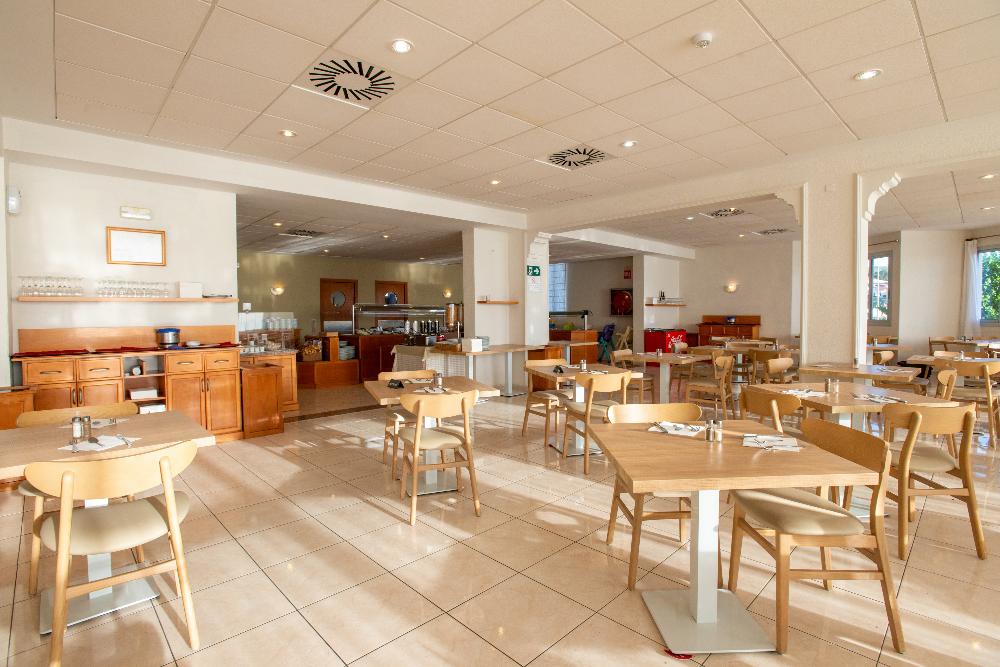 A spacious cafeteria interior features light wooden tables and chairs arranged neatly across a tiled floor. The room is brightly lit with natural light and ceiling fixtures, creating an inviting atmosphere. In the background, a self-service counter and storage units line the wall, with tableware and equipment visible. The overall setting appears clean, modern, and ready to welcome guests.