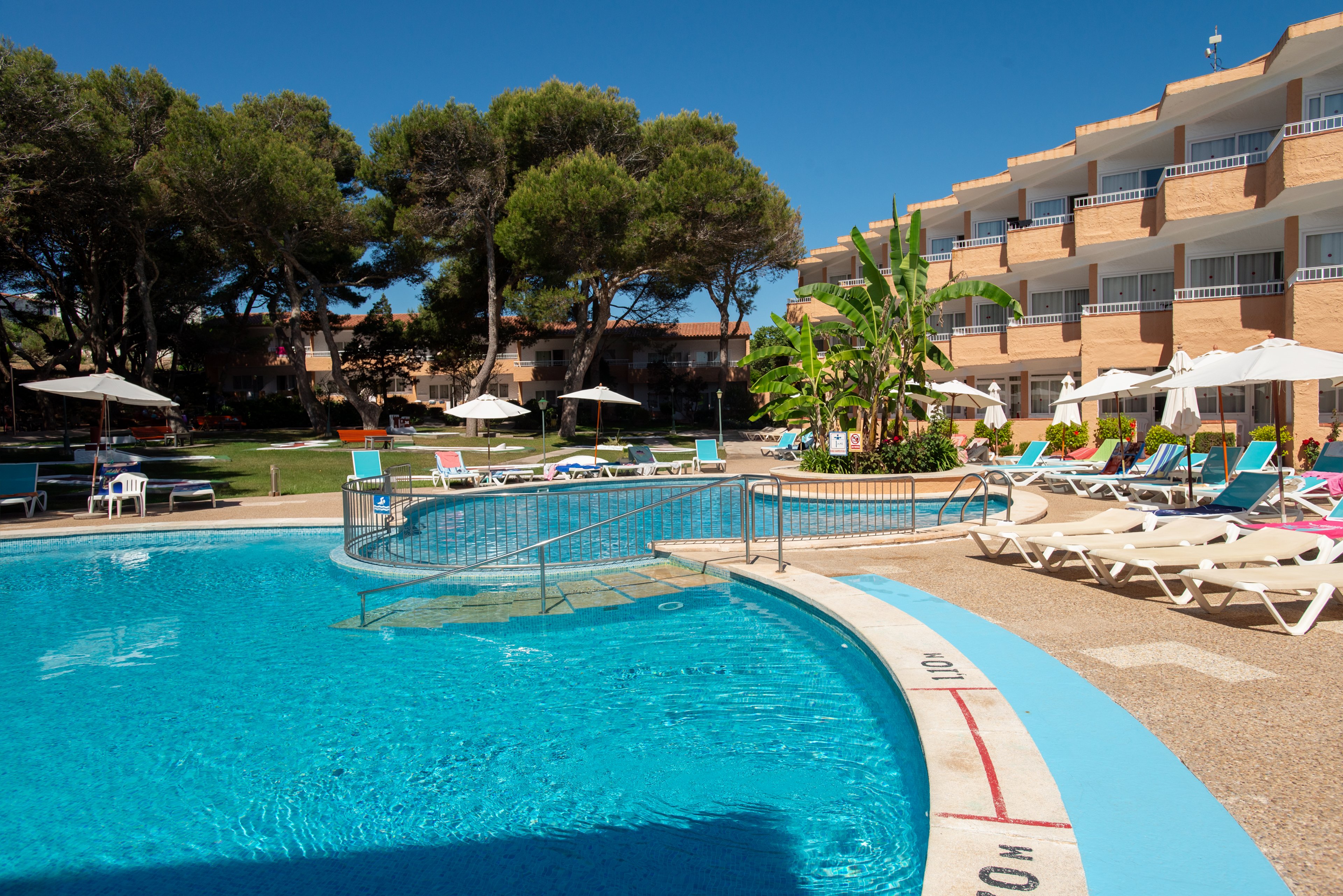 An outdoor swimming pool is surrounded by sun loungers at a hotel resort. The multi-story building with balconies overlooks the pool area and landscaped gardens with trees and tropical plants. Clear blue water and bright sunlight create a relaxing vacation atmosphere. The scene suggests a quiet day at the pool with no people present.