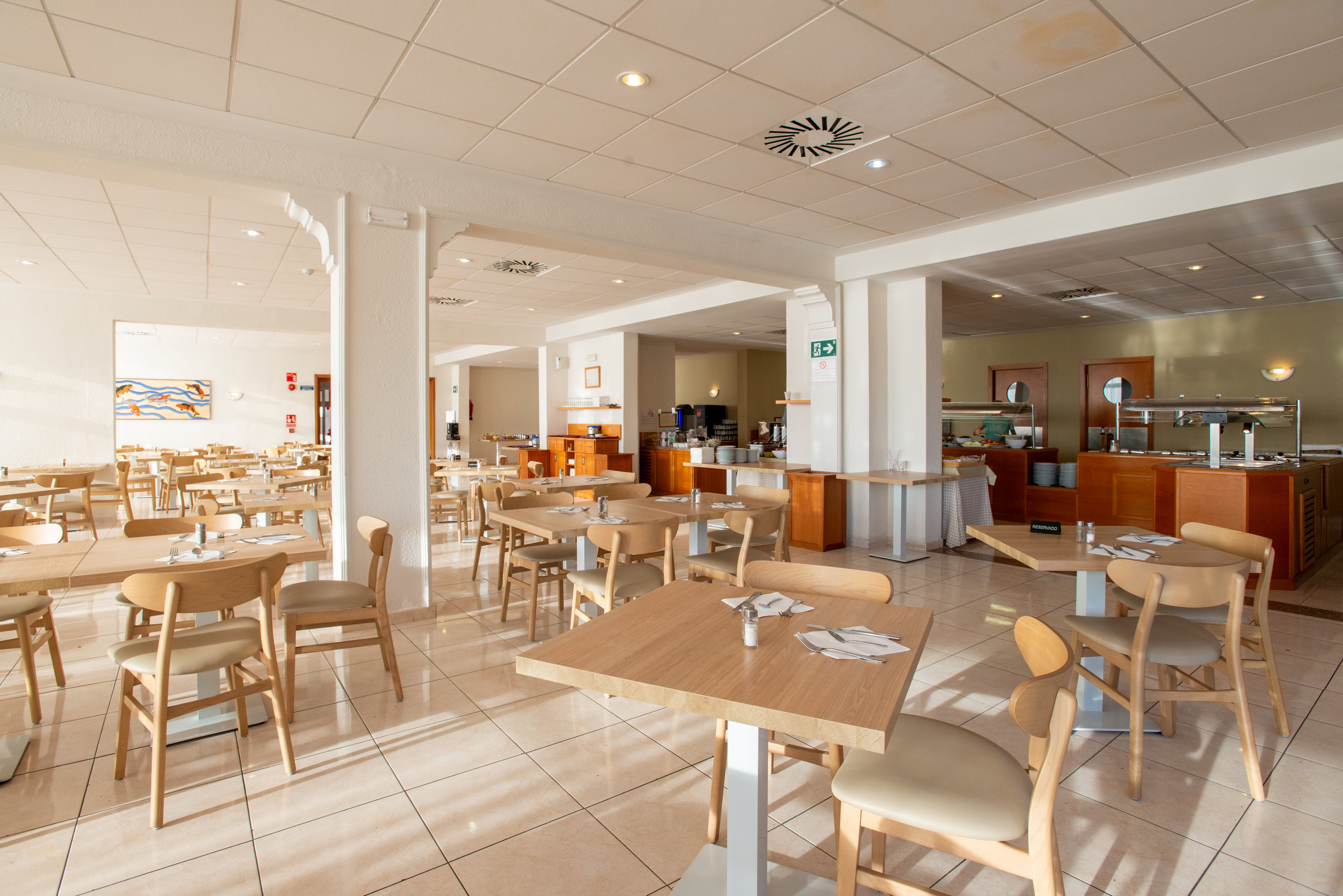 Spacious cafeteria interior with light wooden tables and chairs arranged neatly across a tiled floor. The room is brightly lit with natural and ceiling lighting, creating a clean and welcoming atmosphere. In the background, a self-service buffet area and counters are visible, along with wall art and signage. The overall setting suggests a modern dining hall prepared for guests but currently unoccupied.