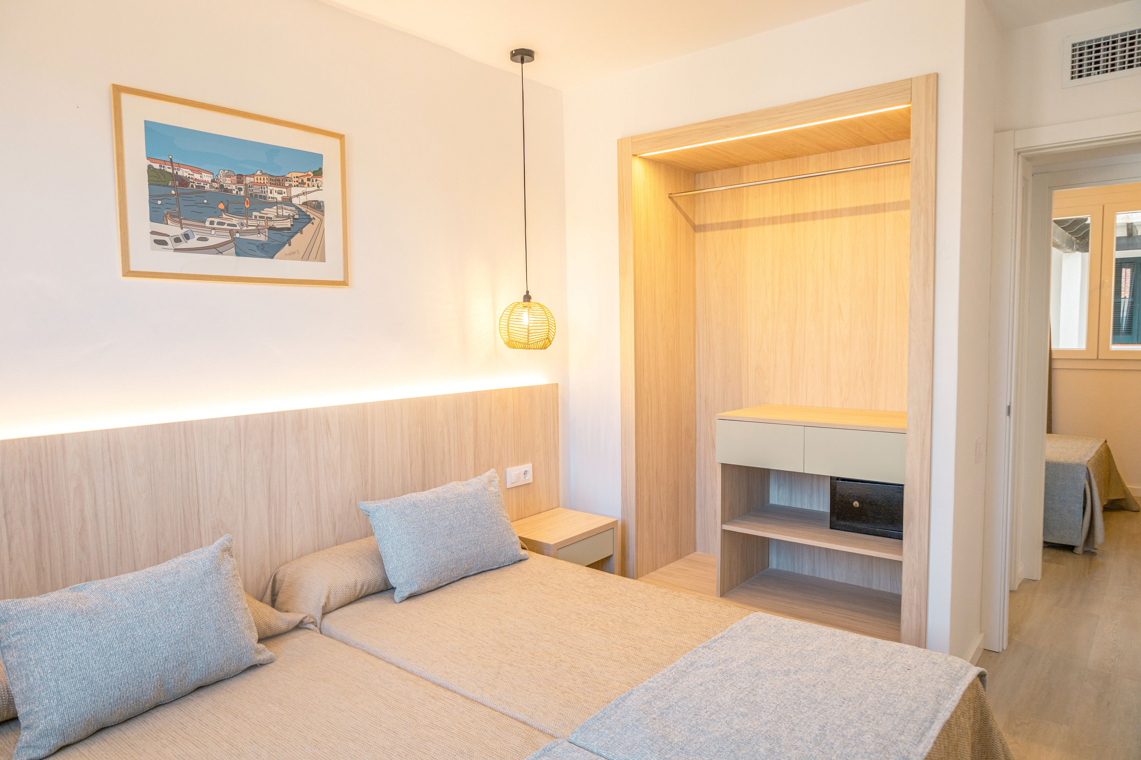 A contemporary hotel room features two neatly made twin beds with light gray bedding and matching pillows. The space is decorated in a minimalist style with pale wood furniture, including an open wardrobe and a small cabinet. Warm ambient lighting from a pendant lamp and integrated strip lights creates a cozy atmosphere. A framed artwork hangs above the headboard, and a doorway leads to an adjacent room or hallway.