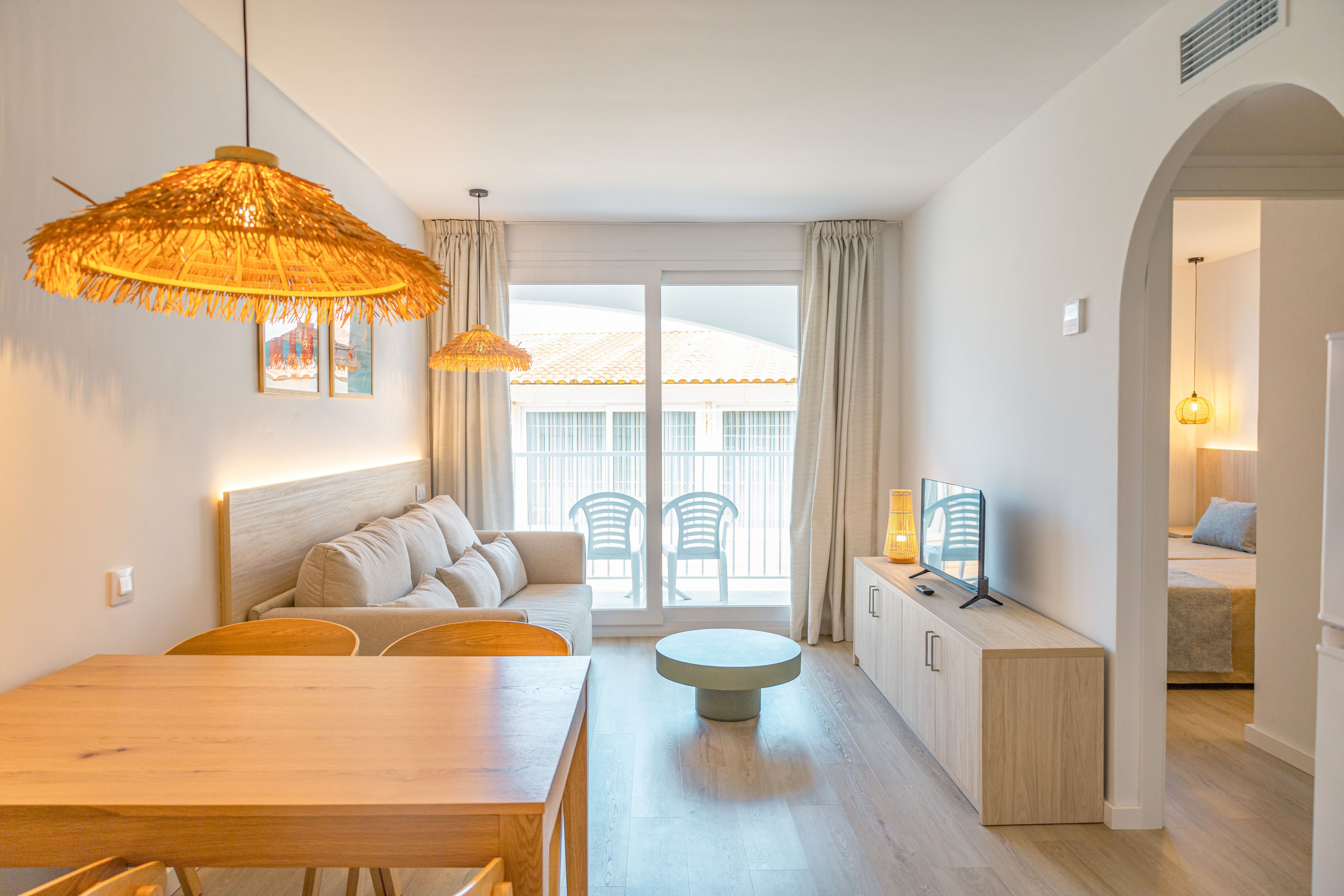 A bright modern living room features a neutral sofa, wooden furniture, and a small round coffee table. Large glass doors open to a balcony with white chairs and a view of tiled rooftops. Warm woven pendant lights and soft curtains create a cozy, coastal-inspired atmosphere. A flat-screen TV sits on a light wood console, with a bedroom visible through an arched doorway.