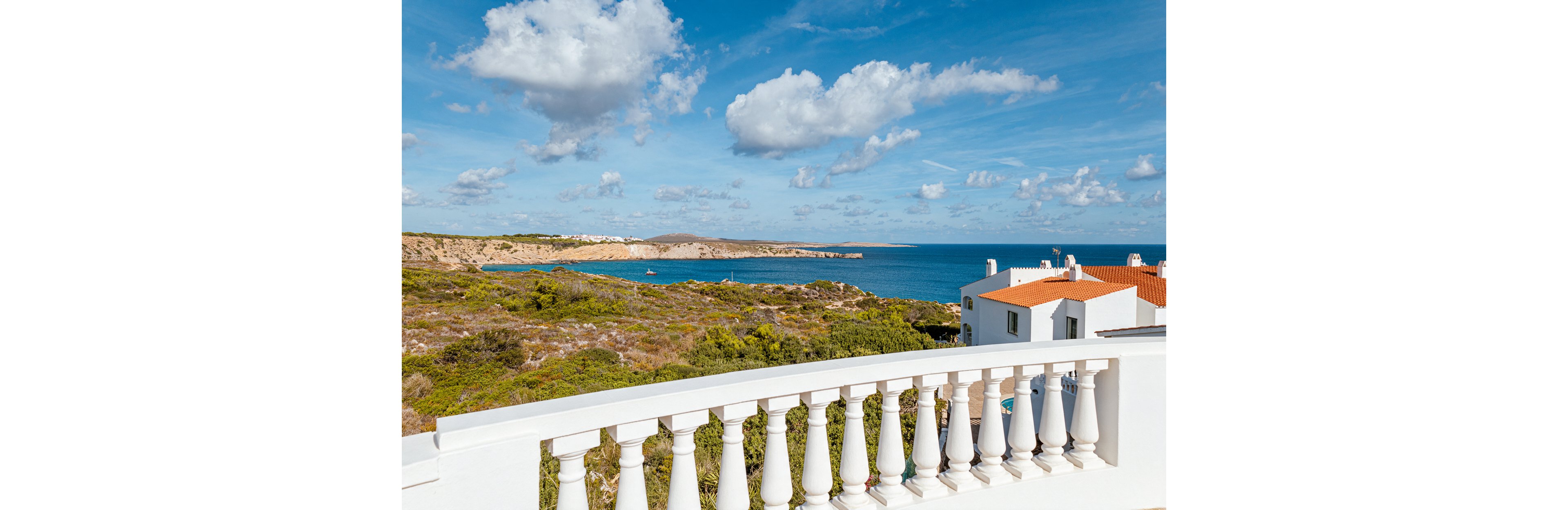 A white balustrade terrace overlooks a rugged coastline and calm blue sea under a bright sky. In the midground, a white house with an orange-tiled roof sits near the cliff edge. The landscape features green shrubs and rocky terrain leading down to the water. Fluffy clouds dot the sky, adding depth and a serene, holiday atmosphere to the scene.