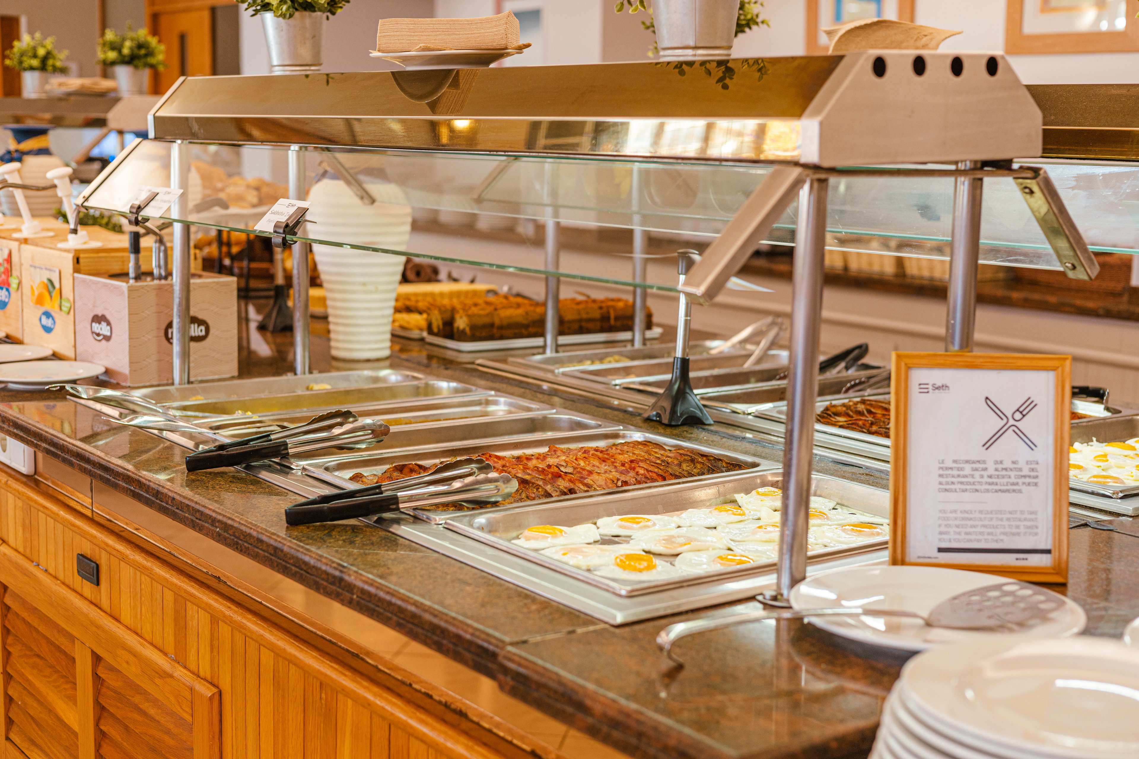 A self-service buffet counter displays a variety of hot dishes in stainless steel trays. Fried eggs, meat in sauce, and other prepared foods are arranged under a glass sneeze guard. Serving tongs and ladles rest on the edge of the pans, ready for guests. In the background are desserts or baked goods, potted plants, and a framed information sign, with stacks of white plates in the foreground.