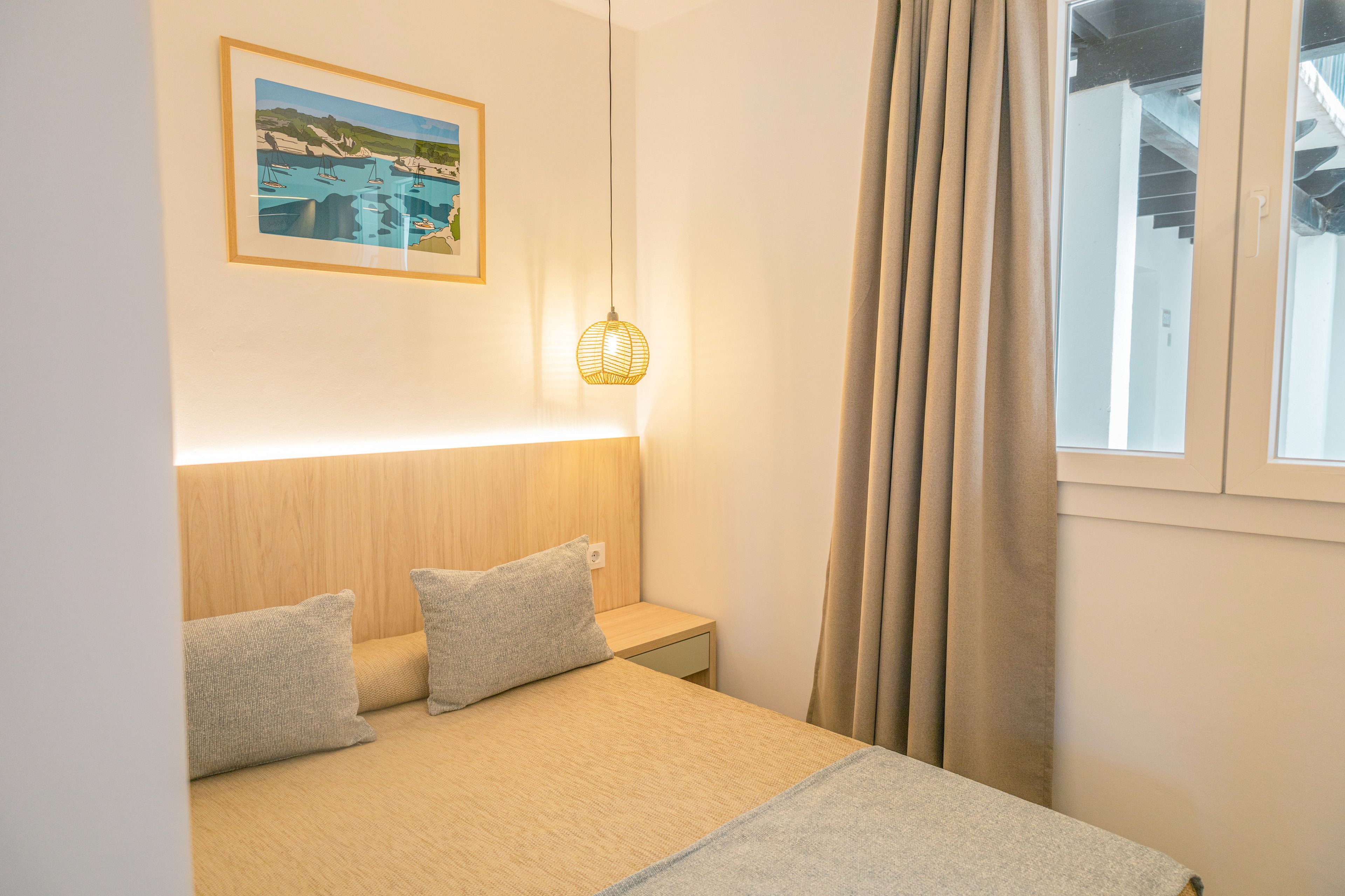 Small contemporary bedroom featuring a neatly made double bed with light beige bedding and two gray cushions. A wooden headboard with integrated strip lighting adds a warm glow to the space. A woven pendant lamp hangs above the bed, complementing the neutral curtains beside a bright window. A framed landscape artwork on the wall introduces a touch of color and visual interest.
