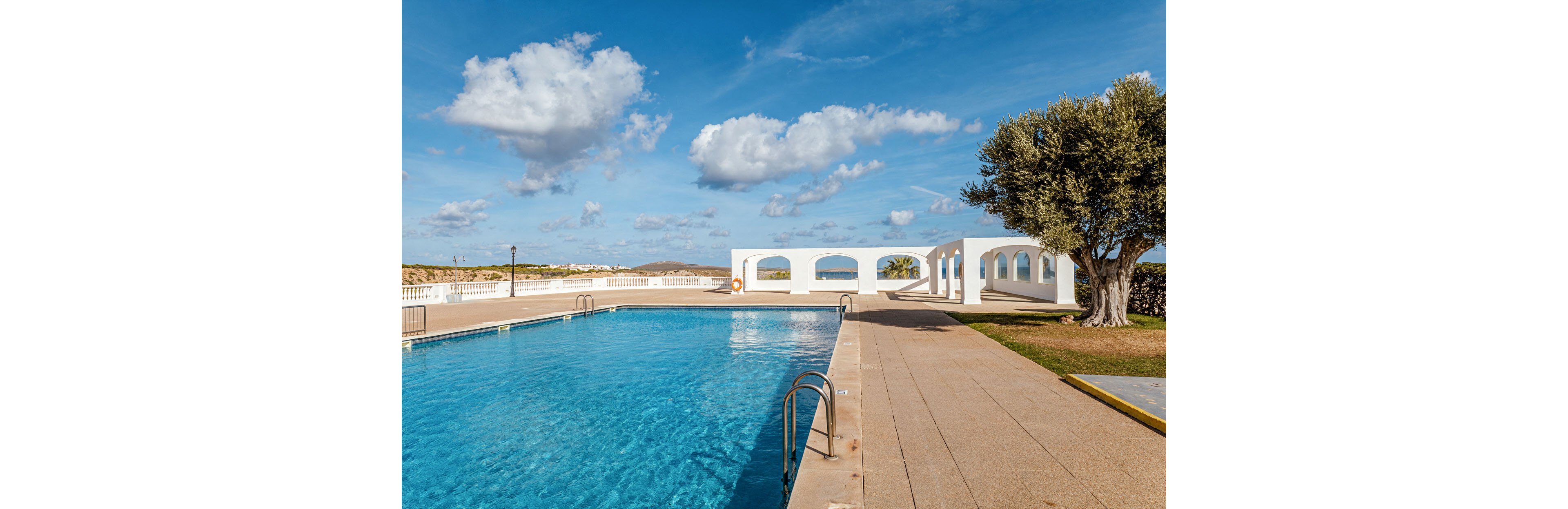 An outdoor swimming pool stretches alongside a paved deck under a bright blue sky. White Mediterranean-style arches frame the background, opening toward a distant coastal landscape. A large tree stands to the right, adding greenery to the sunlit scene. The calm water and scattered clouds create a relaxed vacation atmosphere.