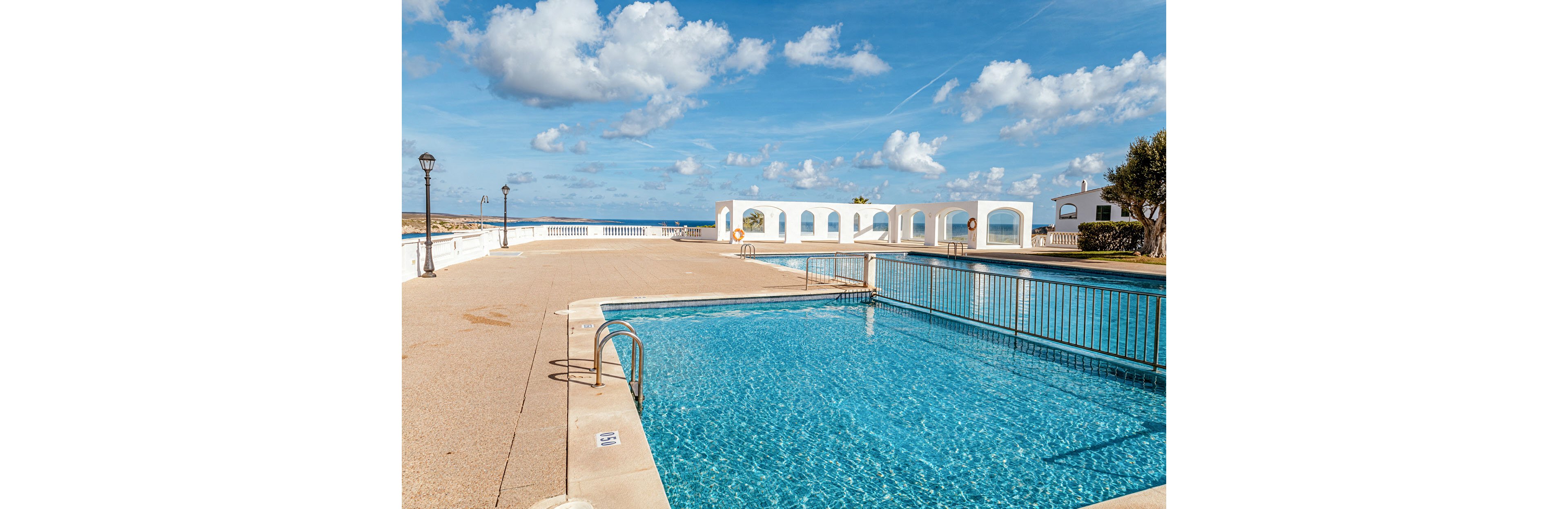 An outdoor swimming pool sits beside a wide paved terrace overlooking the sea. White Mediterranean-style buildings with arches line the background, framed by railings and lampposts. The clear blue water of the pool reflects the bright sky filled with scattered clouds. The scene suggests a quiet, relaxing holiday setting on a sunny day.
