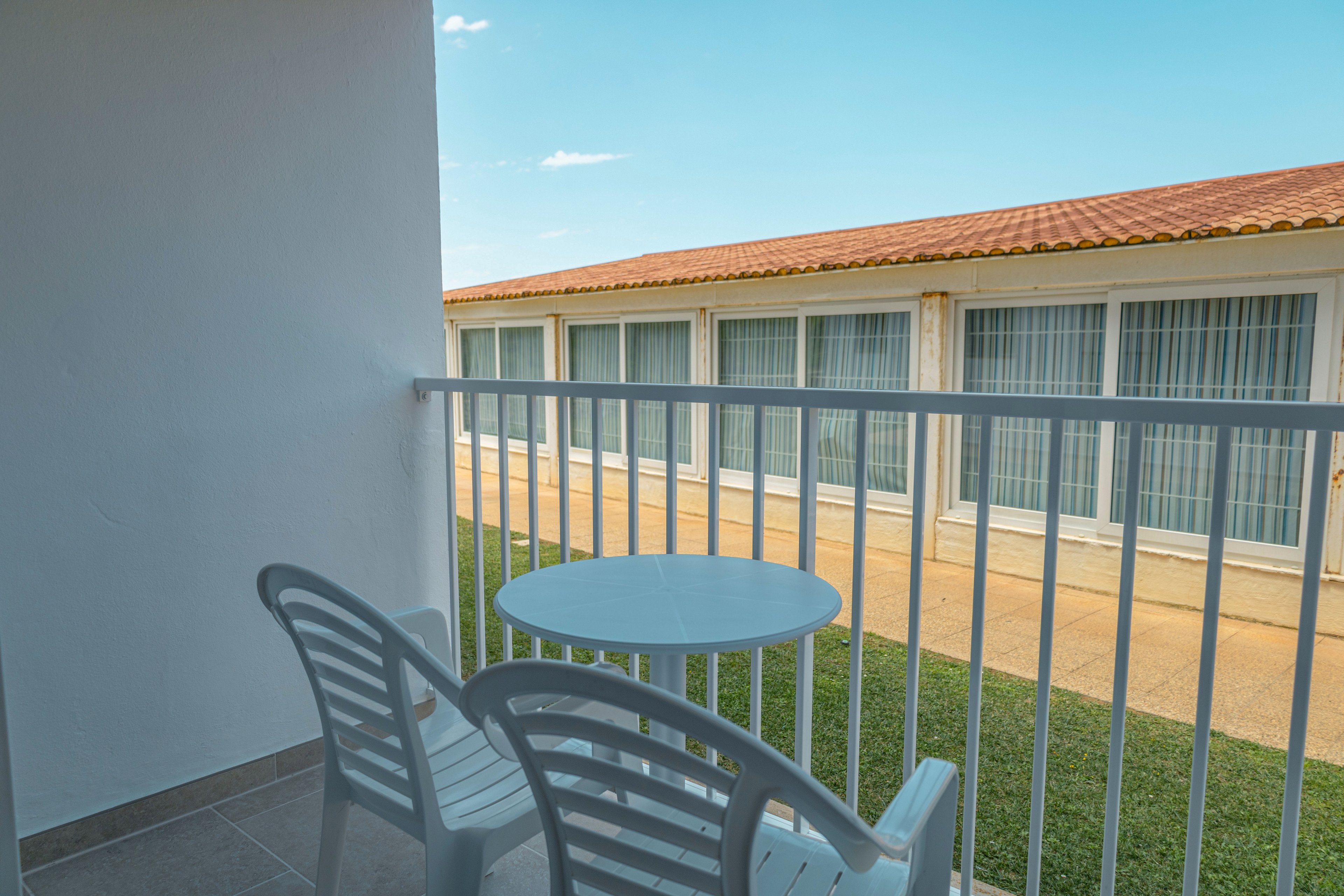 The image shows a small balcony with two white plastic chairs and a round table. A white railing encloses the balcony, overlooking a building with large windows and a tiled roof. The ground below features a strip of green grass and a paved walkway. The scene is lit by clear daylight under a blue sky, suggesting a calm, warm atmosphere.