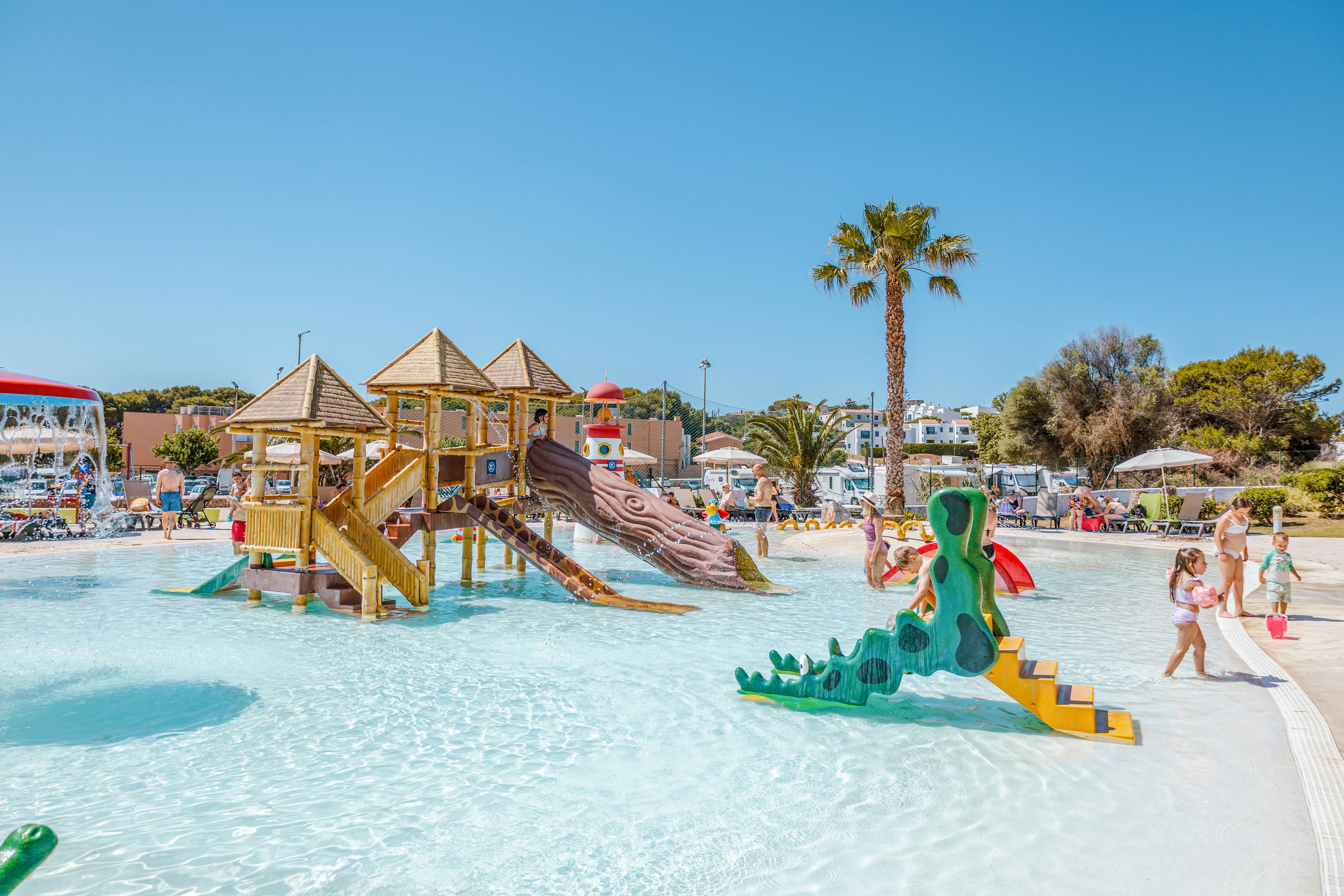 A shallow outdoor pool features a wooden playground structure with multiple slides and climbing areas. Children play in the clear water and walk along the pool edge under a bright blue sky. A green dragon-shaped slide and palm trees add a fun, tropical atmosphere. Resort buildings, sun loungers, and umbrellas are visible in the background, suggesting a family vacation setting.