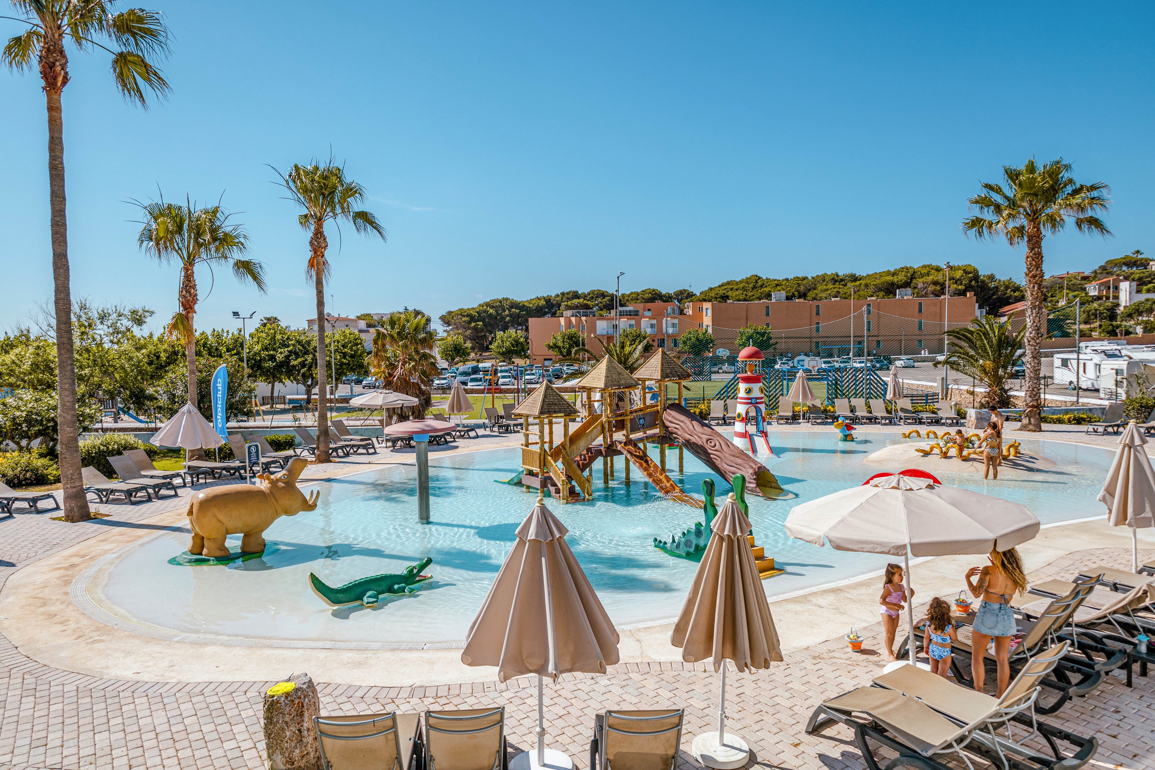 An outdoor family water park features a shallow splash pool with slides and animal sculptures. Children and adults relax and play around the pool under large umbrellas and palm trees. Lounge chairs line the tiled deck, and colorful play structures rise from the water. In the background, parked cars and low buildings indicate a resort or campground setting under a clear blue sky.