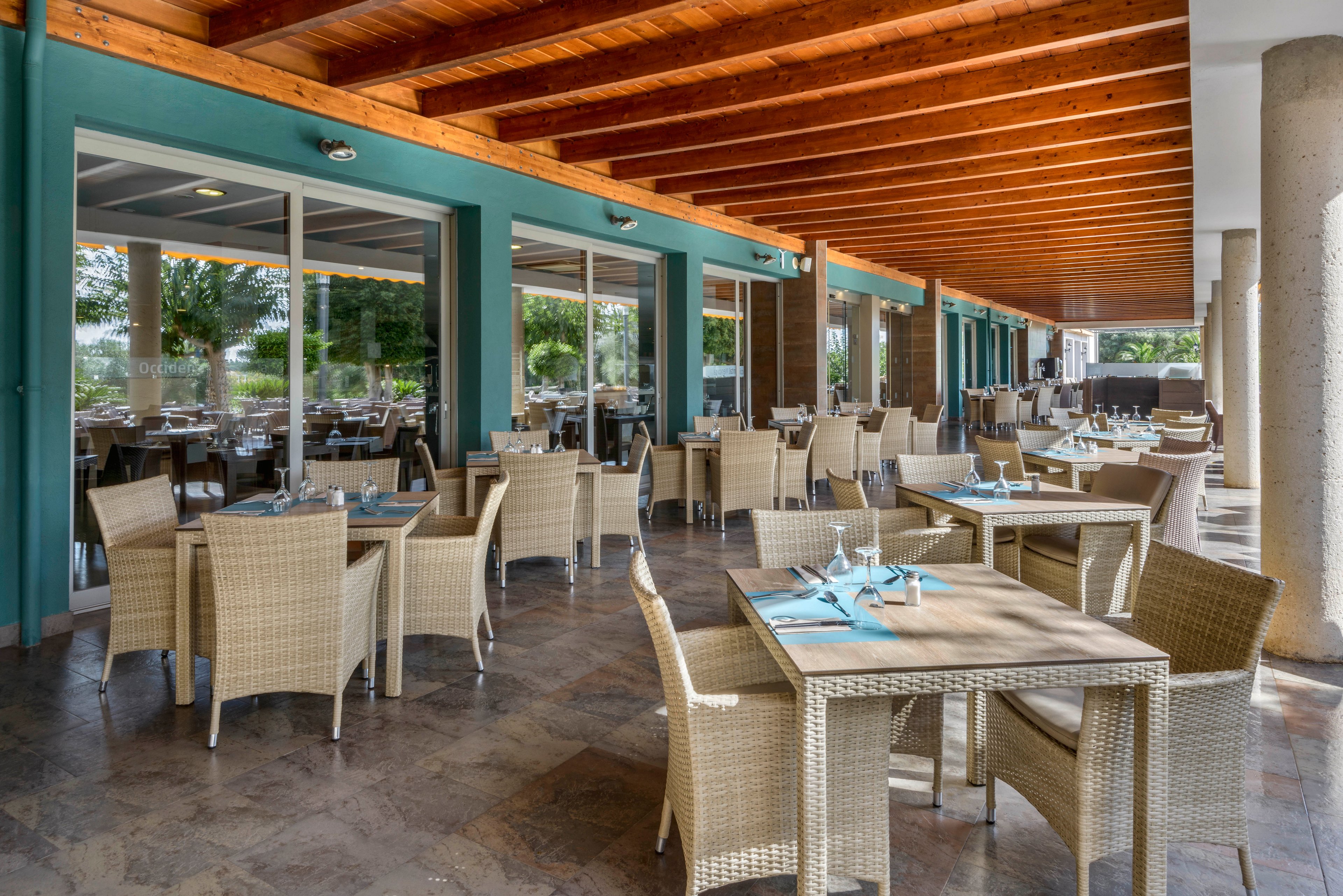 An open-air restaurant terrace features numerous wicker tables and chairs neatly arranged in rows. The space is covered by a wooden beam ceiling and supported by large stone columns, providing shade and structure. Glass doors along one side reflect greenery from the surrounding outdoor area, adding a natural backdrop. Table settings with glasses and napkins suggest a relaxed yet prepared dining environment.