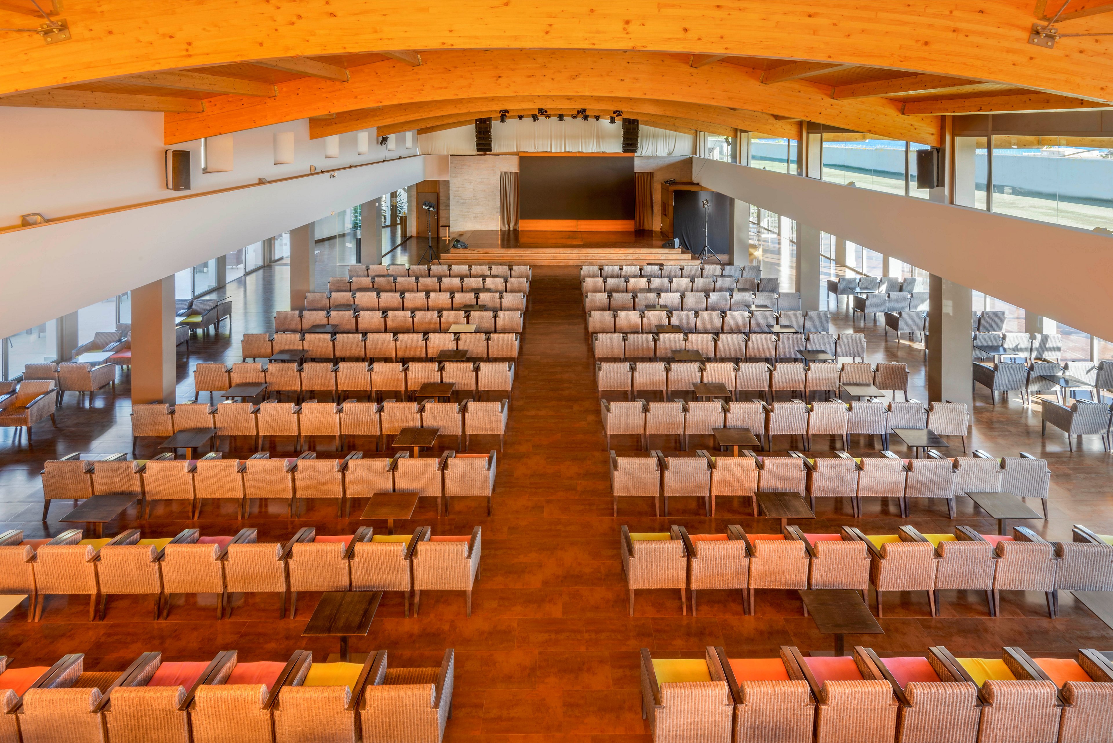 A spacious conference hall is arranged with rows of cushioned chairs facing a central stage. The room is empty, suggesting preparation for a presentation, seminar, or corporate event. Large windows on the side let in natural light and offer views outside. Warm wooden beams and flooring create a modern yet inviting atmosphere.