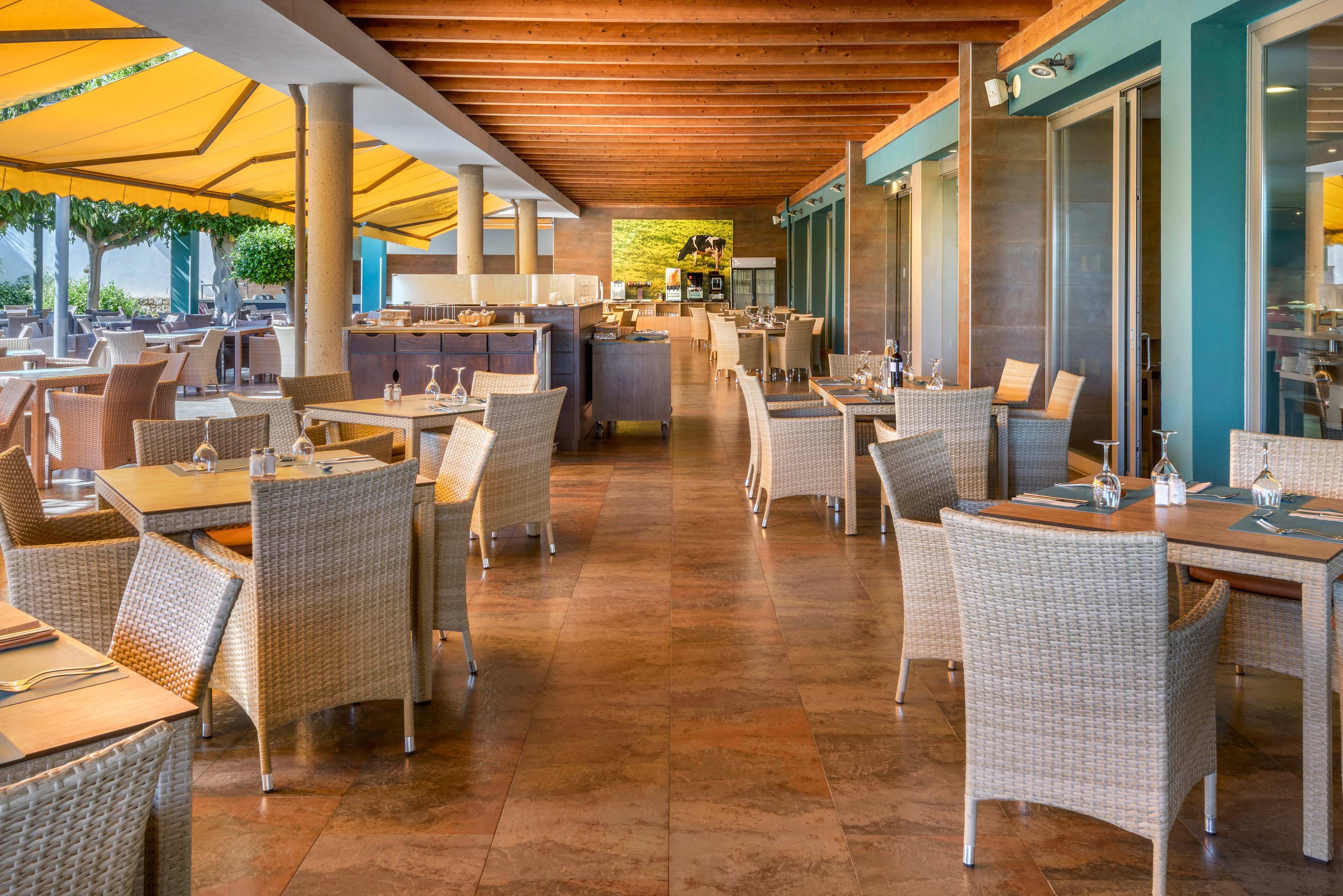 An open-air restaurant terrace features neatly arranged wicker tables and chairs. The space is covered by a wooden ceiling and large yellow awnings that provide shade. Glass doors line one side, leading into the interior of the building. The setting appears calm and inviting, with tableware placed and ready for guests.