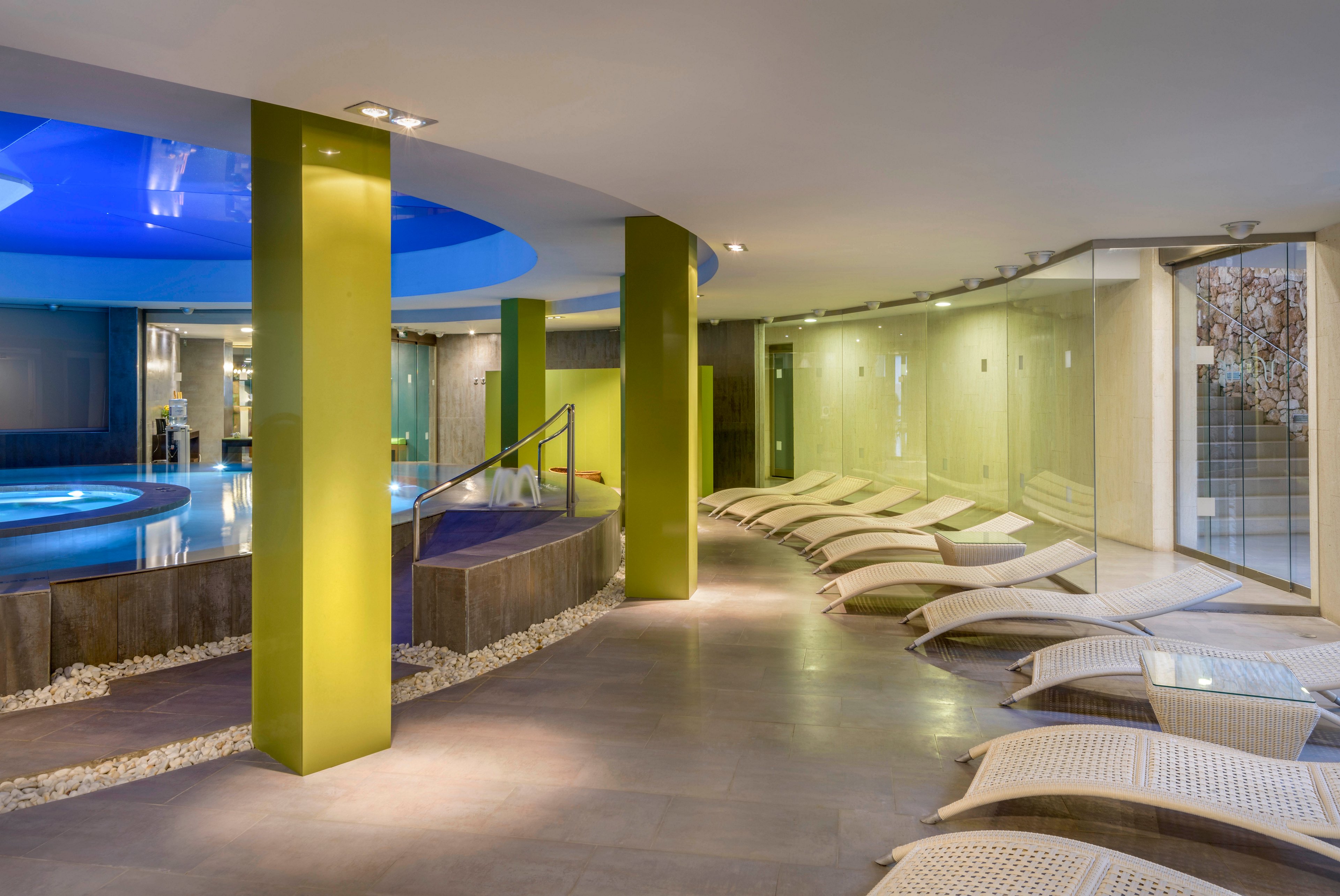 A contemporary indoor spa features a circular pool surrounded by sleek green columns. Curved white lounge chairs line the tiled floor, creating a relaxing seating area. Glass walls and soft recessed lighting enhance the clean, minimalist design. The blue-lit ceiling above the pool adds a tranquil, luxurious atmosphere.