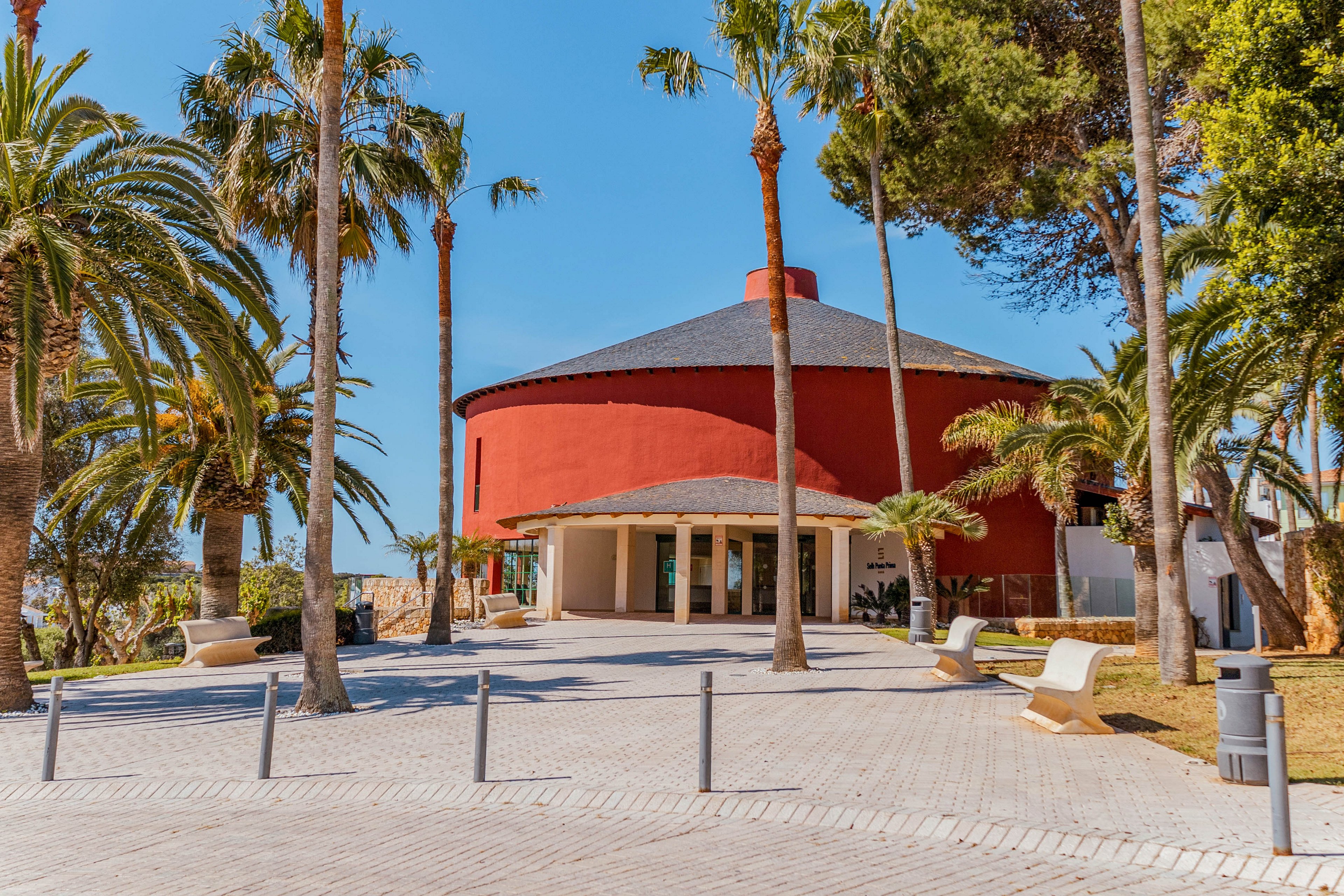 A distinctive red circular building stands in a sunny outdoor plaza lined with palm trees. The structure features a low, sloping roof and a shaded entrance supported by columns. Bright daylight and clear blue skies create a warm, inviting atmosphere. Benches, bollards, and a paved walkway lead toward the building, suggesting a public or cultural space. No people are visible in the scene, emphasizing the architecture and landscape.