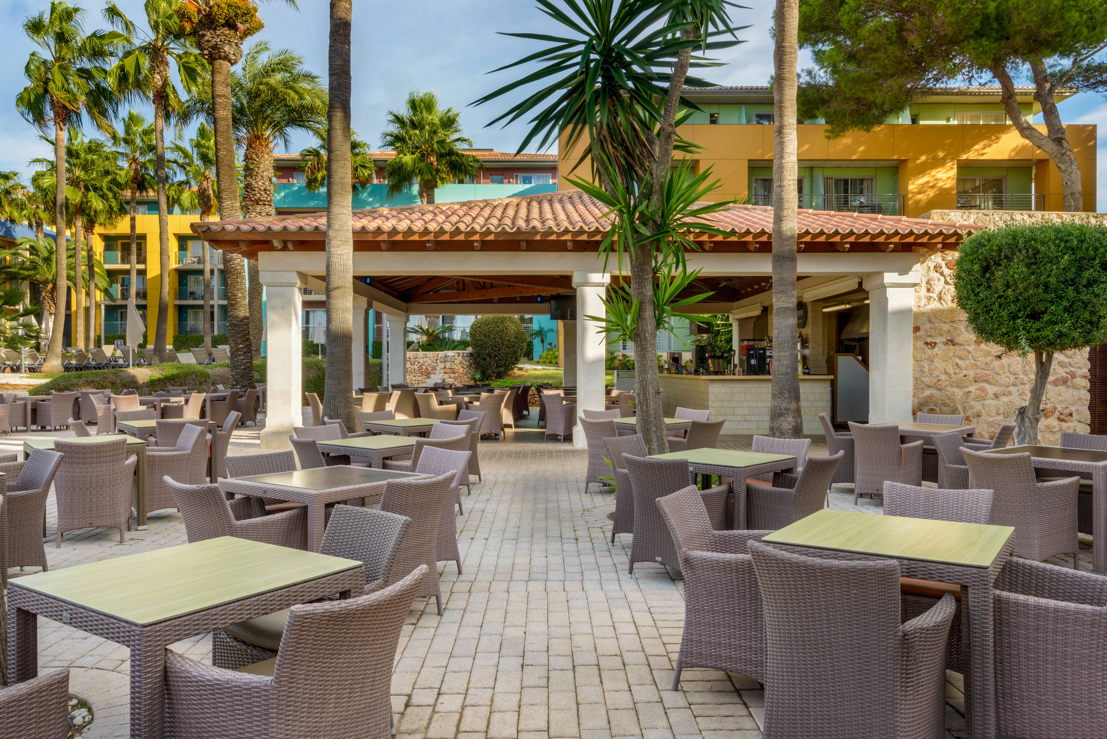 An open-air resort cafe features numerous empty tables and wicker chairs arranged on a tiled patio. The setting is surrounded by tall palm trees and lush greenery, creating a relaxed vacation atmosphere. A covered bar area stands at the center with a terracotta roof and stone accents. Colorful hotel buildings in the background add to the warm, Mediterranean-style ambiance.