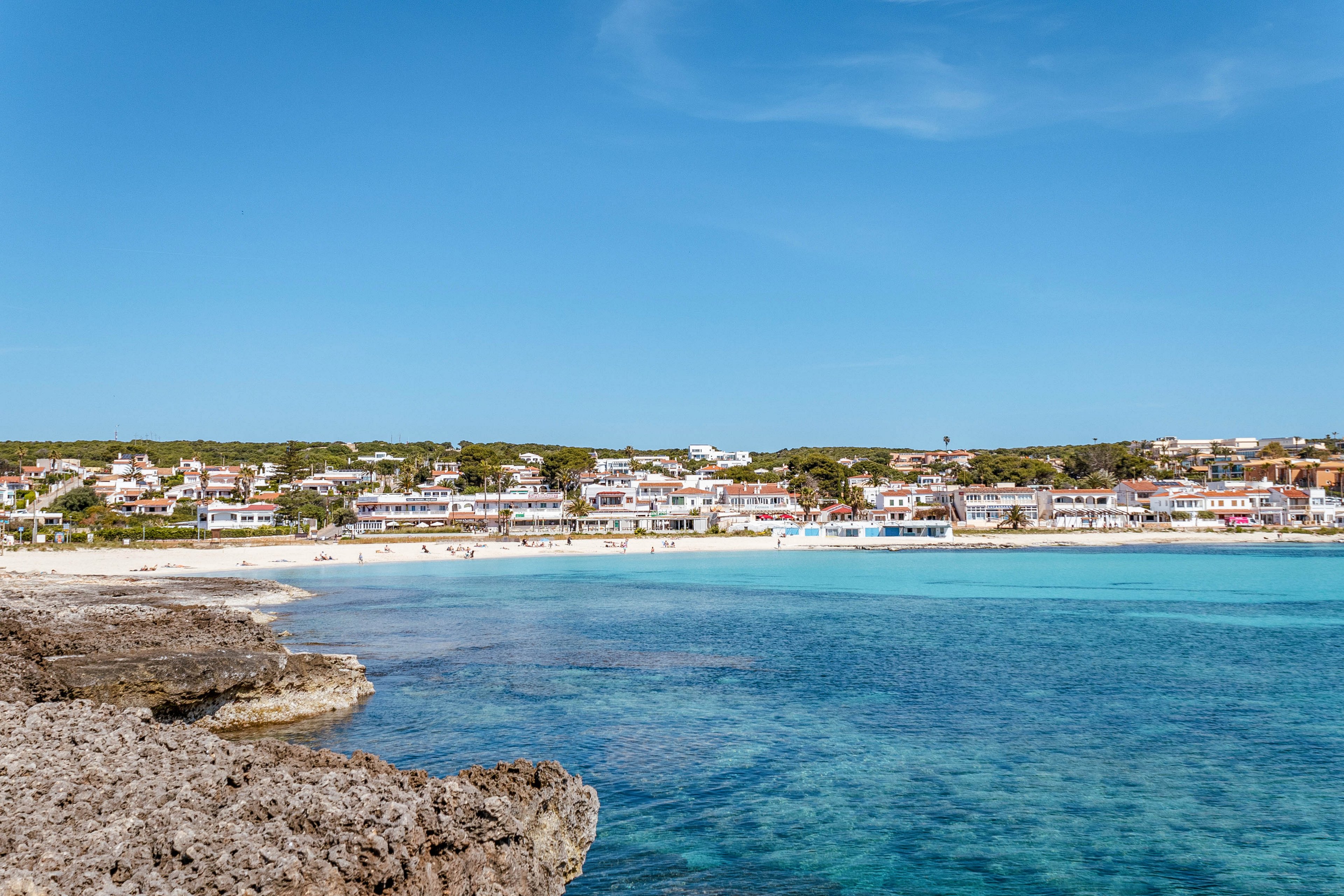 A picturesque coastal town lines a bright turquoise bay under a clear blue sky. Low-rise houses and buildings stretch along the sandy shoreline in the distance. Rocky outcrops frame the foreground, contrasting with the calm, transparent water. The scene suggests a peaceful seaside destination ideal for relaxation and swimming.