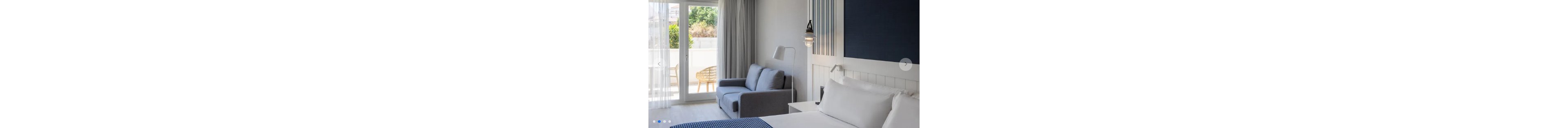 A contemporary hotel room features a neatly made bed with white linens and a blue accent blanket. Beside the bed, a gray sofa and floor lamp sit near large glass doors leading to a sunny balcony. Sheer curtains soften the natural light, revealing outdoor wicker chairs and greenery beyond. The overall design is minimal, airy, and inviting with a cool blue and neutral color palette.