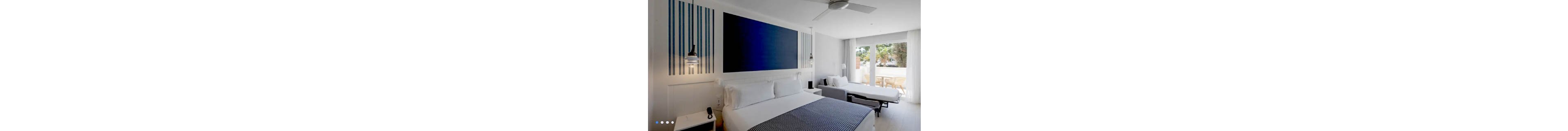 A contemporary hotel room features a large bed in the foreground with white linens and a blue patterned throw. A second bed or sofa bed is positioned near the sliding glass doors that open to a balcony with outdoor chairs and greenery visible outside. The interior decor is minimalist, with white walls, blue striped accents, and a large dark blue wall panel above the main bed. Natural light floods the space, creating an airy and inviting atmosphere.
