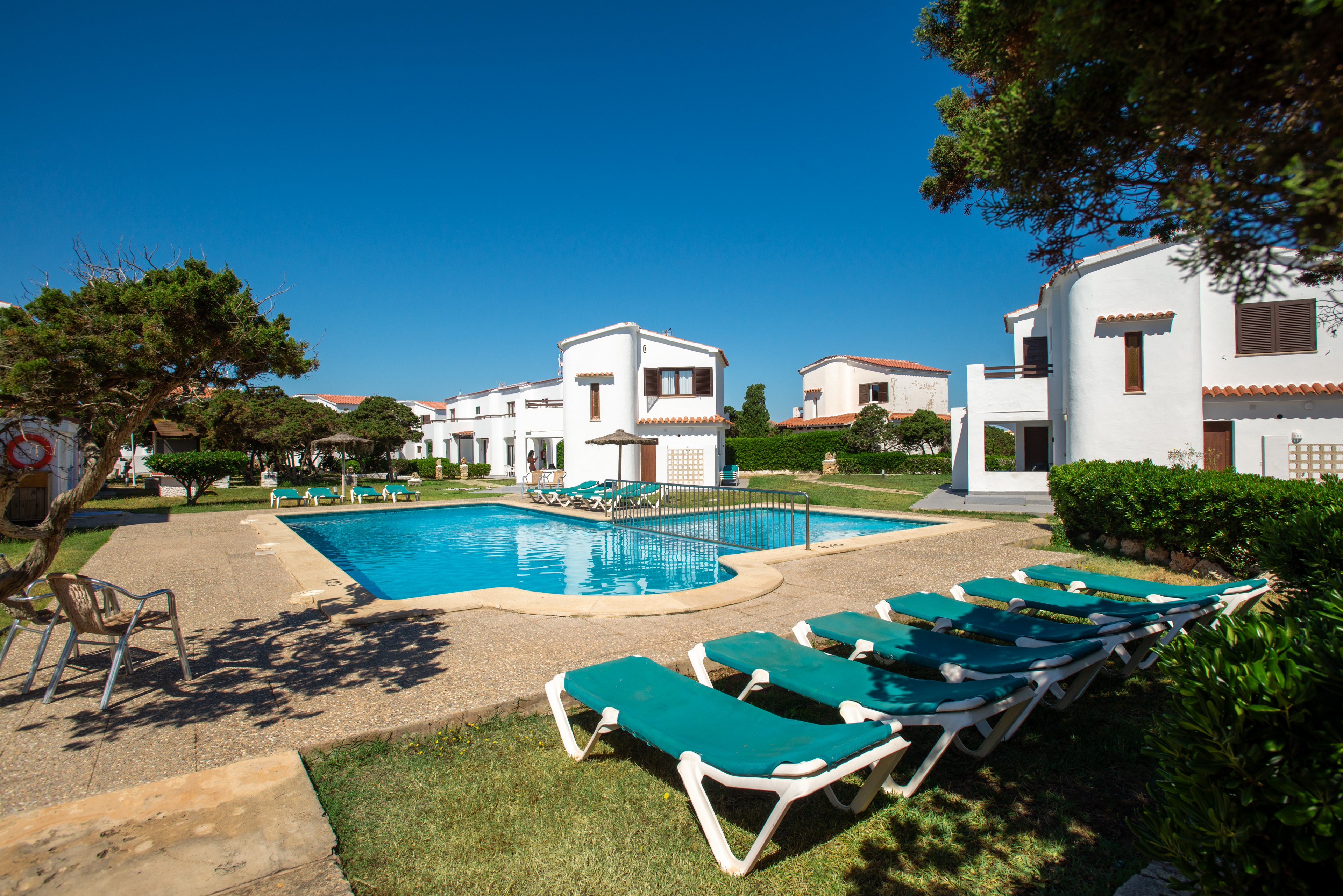 Outdoor swimming pool surrounded by green sun loungers in a sunny resort setting. White two-story villas with terracotta roofs frame the pool area, with manicured lawns and pathways connecting the buildings. Clear blue sky and bright daylight create a warm, inviting vacation atmosphere. Trees and shrubs add natural greenery around the leisure space.