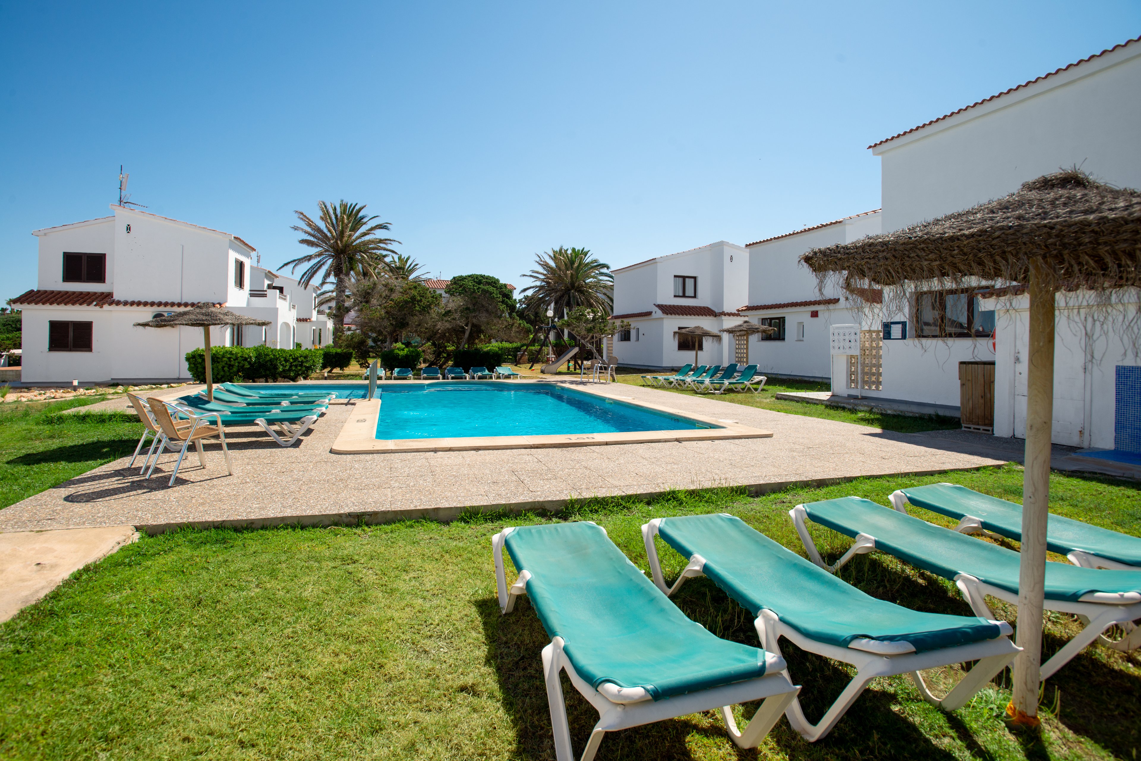 A rectangular outdoor swimming pool is surrounded by sun loungers in a grassy courtyard between white holiday villas. Palm trees and a clear blue sky create a bright, summery atmosphere. The scene suggests a quiet vacation resort with space for relaxation by the water. No people are present, emphasizing the tranquil and inviting setting.