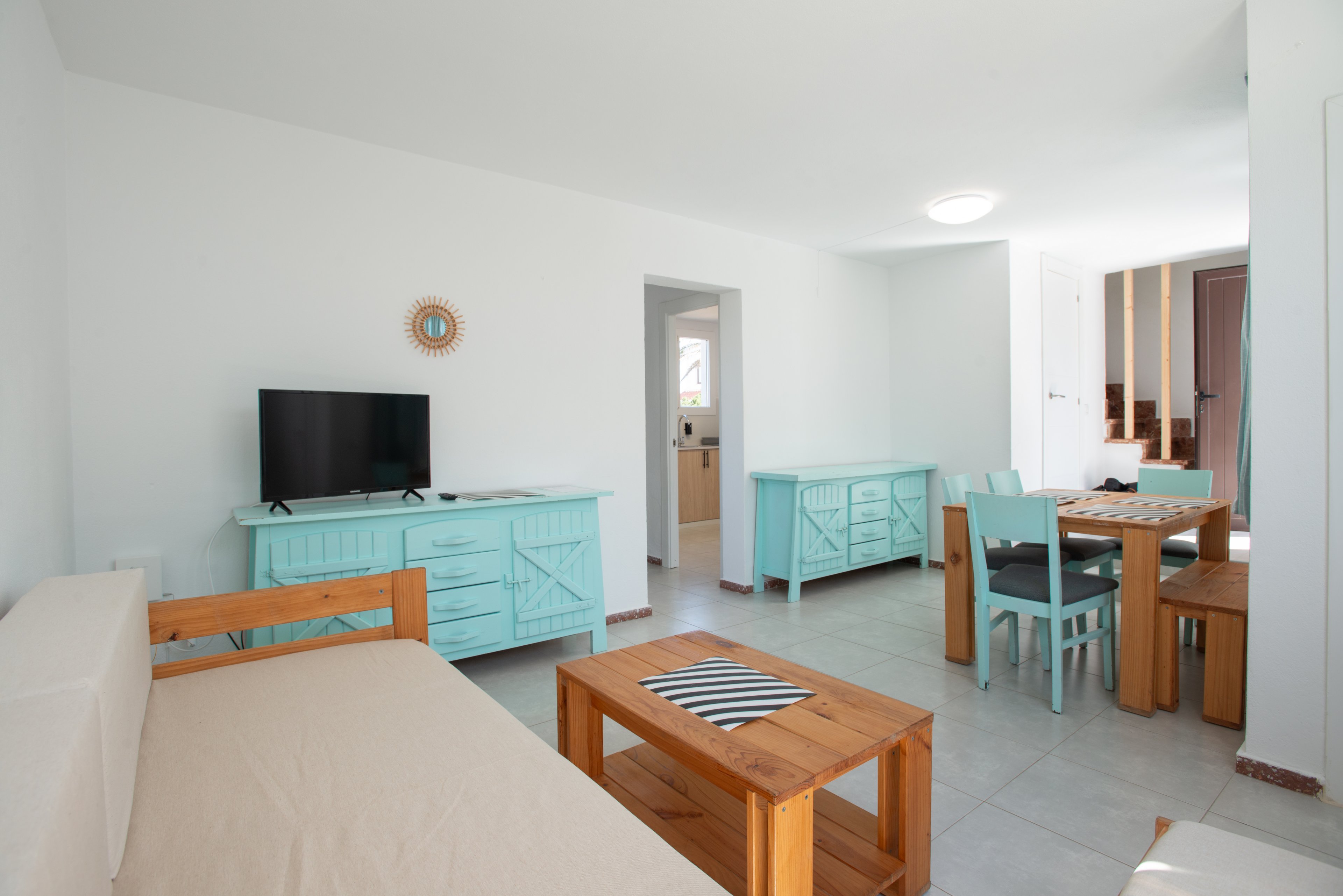 A bright living room features light turquoise cabinets and dining chairs paired with natural wood furniture. A flat-screen television sits on a turquoise sideboard against a white wall, with a small decorative mirror above it. The space includes a wooden dining table, coffee table, and sofa with neutral cushions, creating a relaxed coastal feel. Doorways lead to adjacent rooms, adding depth and openness to the interior layout.