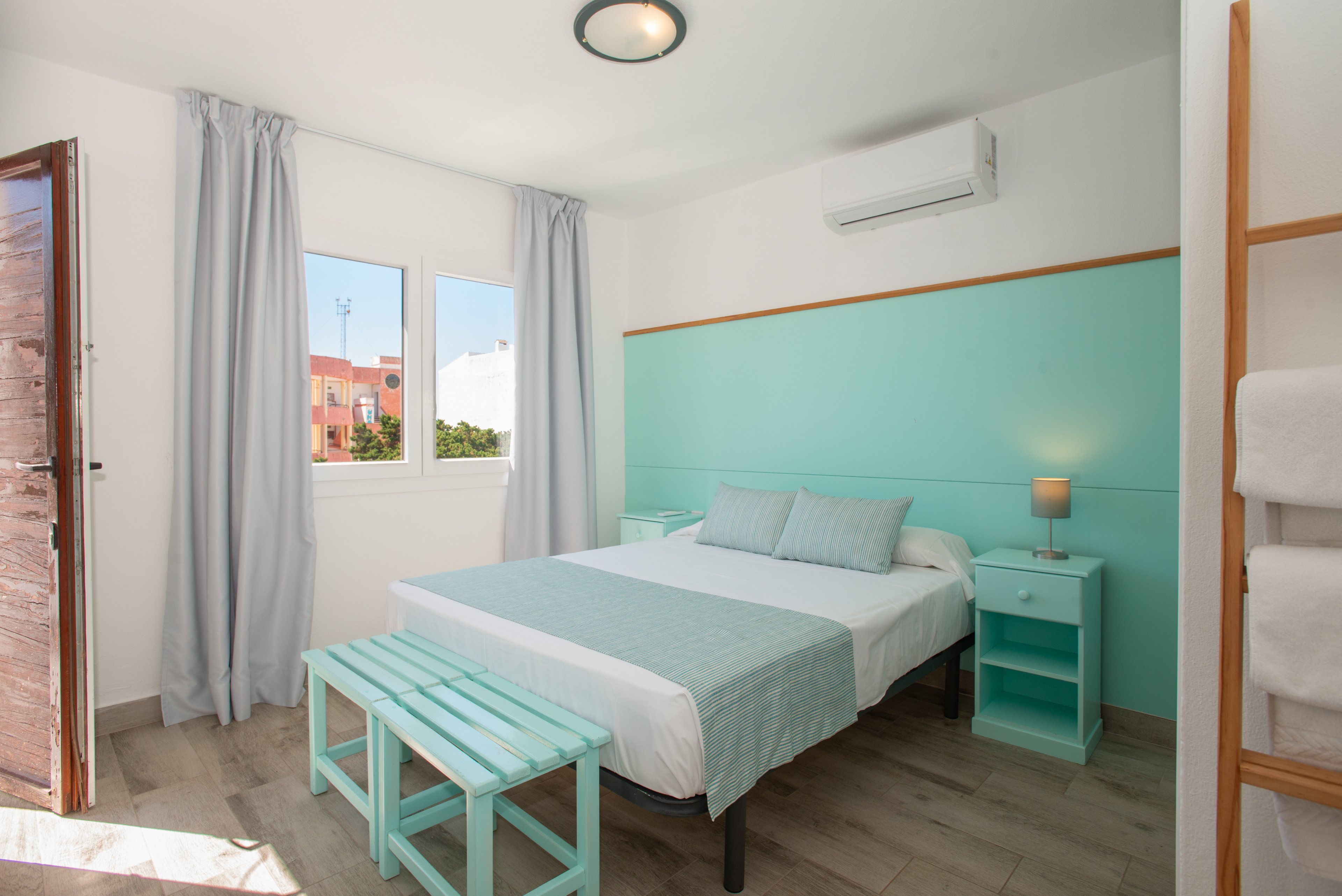 A sunlit bedroom features a double bed with mint-colored headboard, bench, and bedside tables. Light gray curtains frame the window, revealing a view of neighboring buildings and greenery. A small lamp on the nightstand adds warmth, while an air conditioning unit is mounted above the bed. The room has light wood flooring and a clean, minimalist decor that feels fresh and inviting.