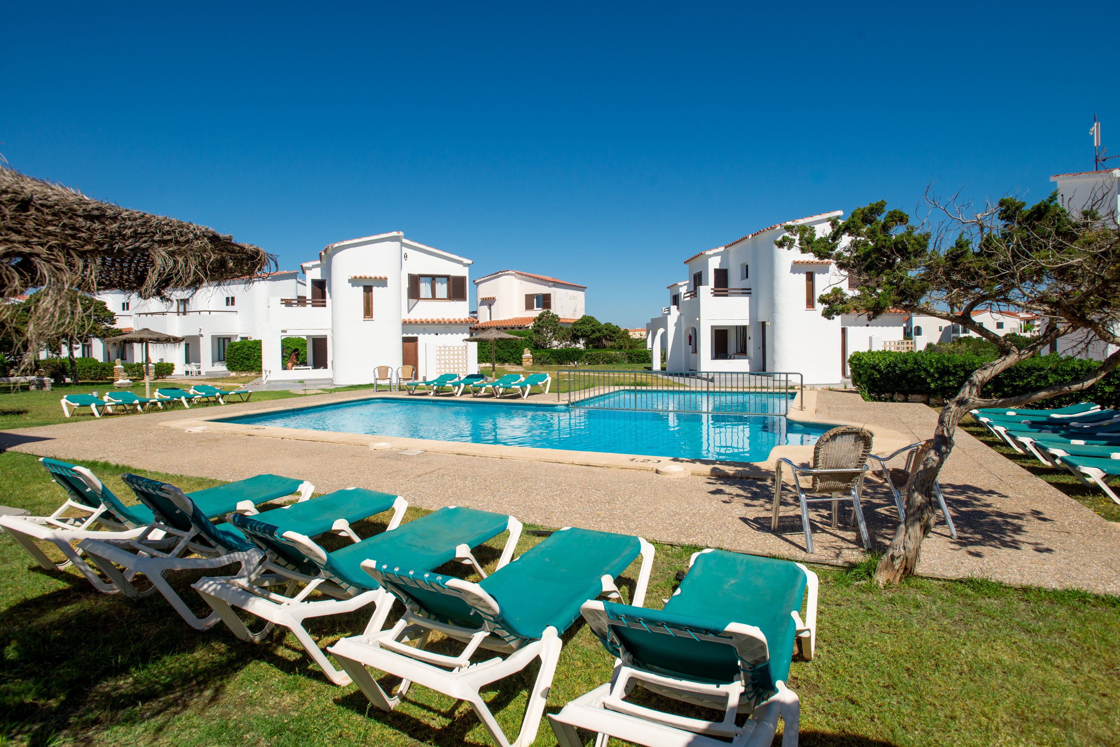 An outdoor swimming pool is surrounded by green sun loungers in a sunny resort setting. White low-rise buildings with balconies frame the pool area, creating a clean Mediterranean-style atmosphere. The sky is clear and blue, and the grass and shrubs add a touch of greenery around the paved pool deck. A few empty chairs and a shaded structure suggest a quiet, relaxing vacation environment.