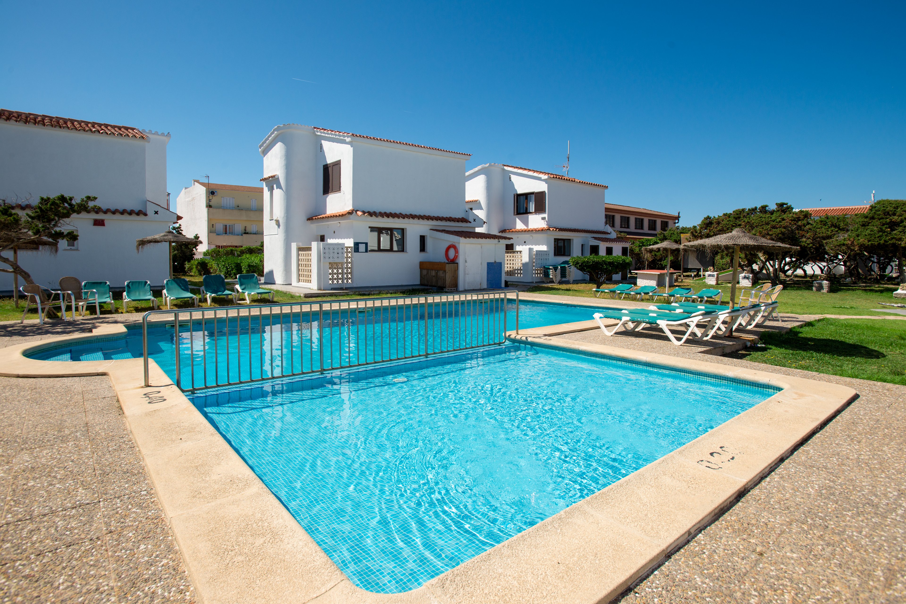A clear blue swimming pool sits in front of white holiday villas under a bright, cloudless sky. Sun loungers and parasols are arranged around the pool on green lawns, suggesting a relaxing vacation setting. The architecture features simple, white facades with terracotta roof tiles and multiple windows. The overall scene conveys a peaceful, sunny resort atmosphere ideal for leisure and family stays.