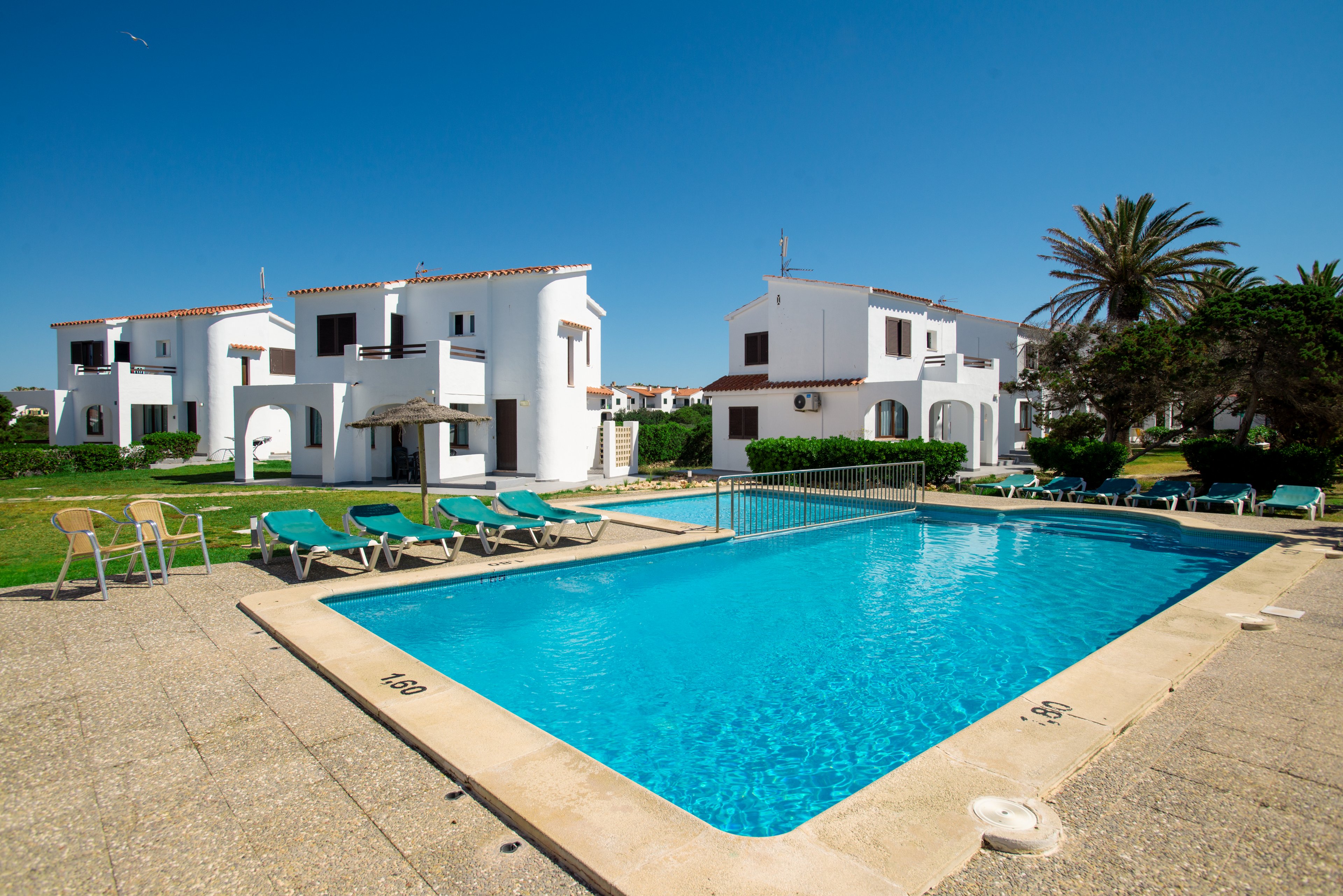 An outdoor swimming pool sits at the center of a sunny resort, surrounded by green lawns and white Mediterranean-style villas. Several sun loungers line the poolside terrace, with a few chairs and a small table nearby. Palm trees and manicured shrubs add a tropical feel under a clear blue sky. The scene suggests a quiet vacation setting ideal for relaxation and leisure.