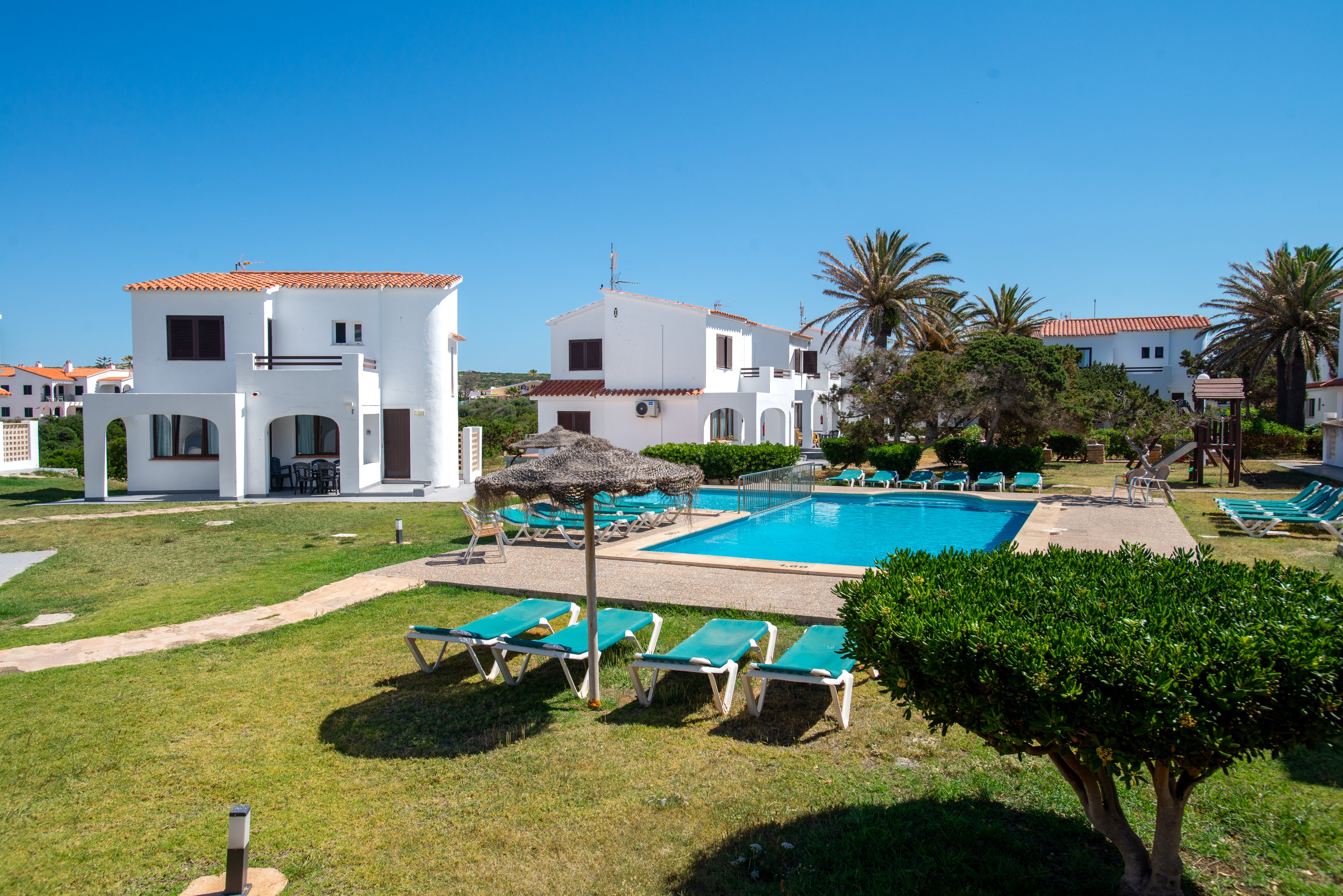 Image of a small white villa complex surrounding an outdoor swimming pool under a clear blue sky. Sun loungers and straw parasols are arranged around the pool on green lawns and paved paths. Palm trees and trimmed bushes add a tropical, vacation feel to the scene. The bright daylight and empty loungers suggest a peaceful, relaxing holiday setting.
