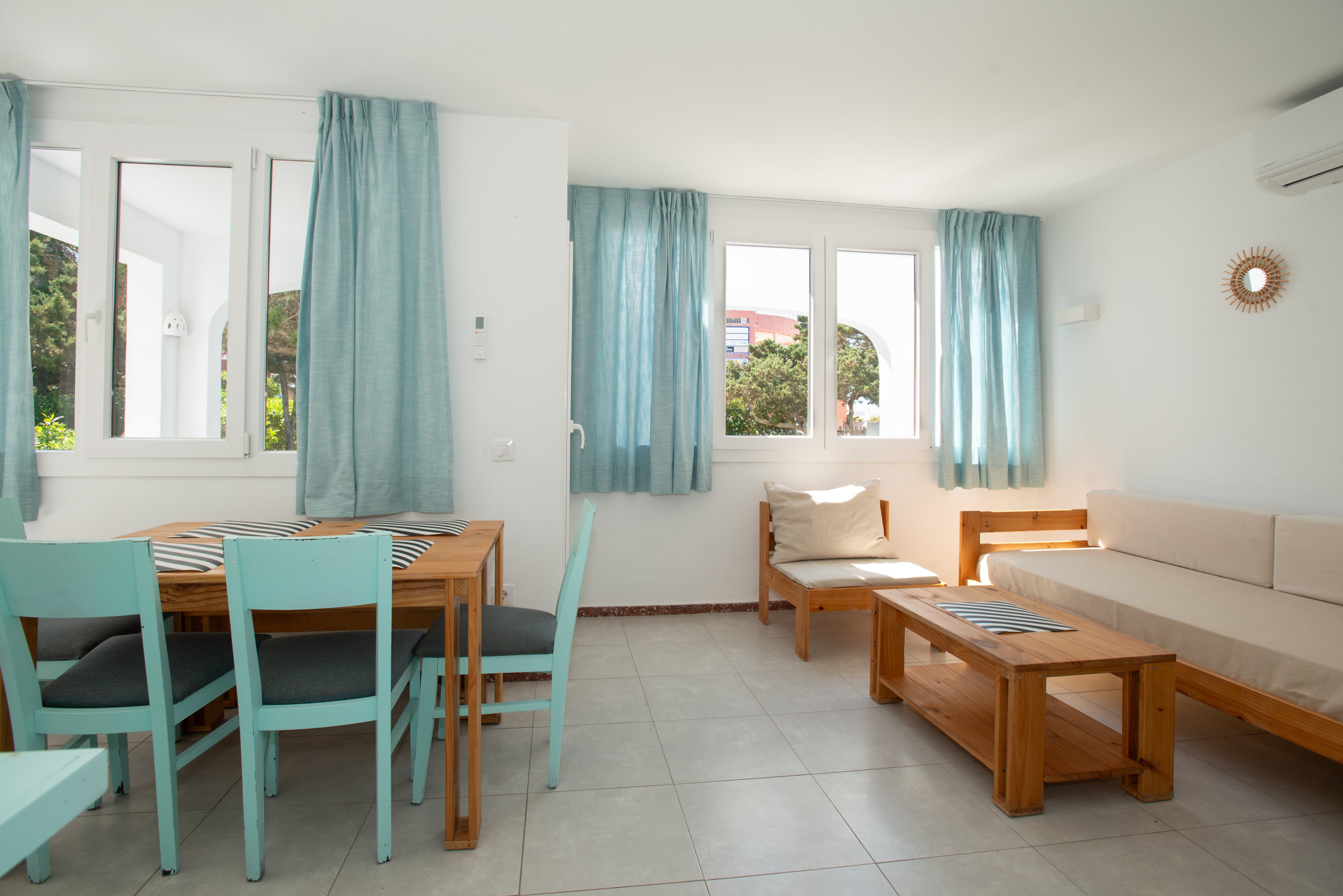 Sunlit living and dining area features light wooden furniture and soft beige upholstery. Turquoise curtains and matching dining chairs add a coastal touch to the white-walled room. Large windows let in natural light and offer views of greenery and distant buildings. The space feels clean, minimal, and inviting, with neutral tiled flooring and simple decor.