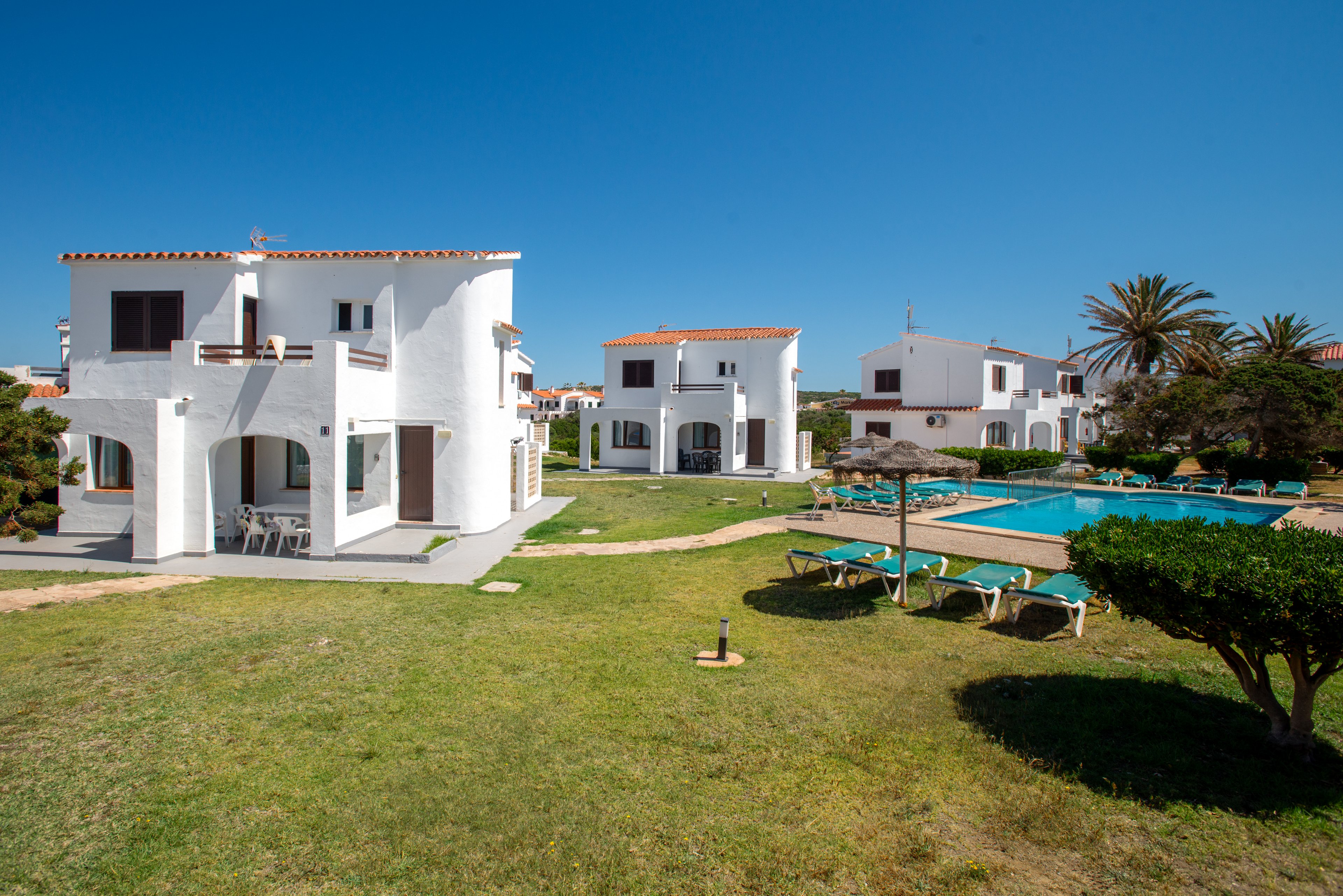 White two-story villas stand around a grassy courtyard with a bright blue outdoor swimming pool. Sun loungers are arranged beside the pool, suggesting a relaxing holiday or resort setting. The sky is clear and blue, and palm trees and trimmed shrubs add to the coastal atmosphere. The scene appears quiet and sunny, ideal for vacation rentals or leisure travel imagery.