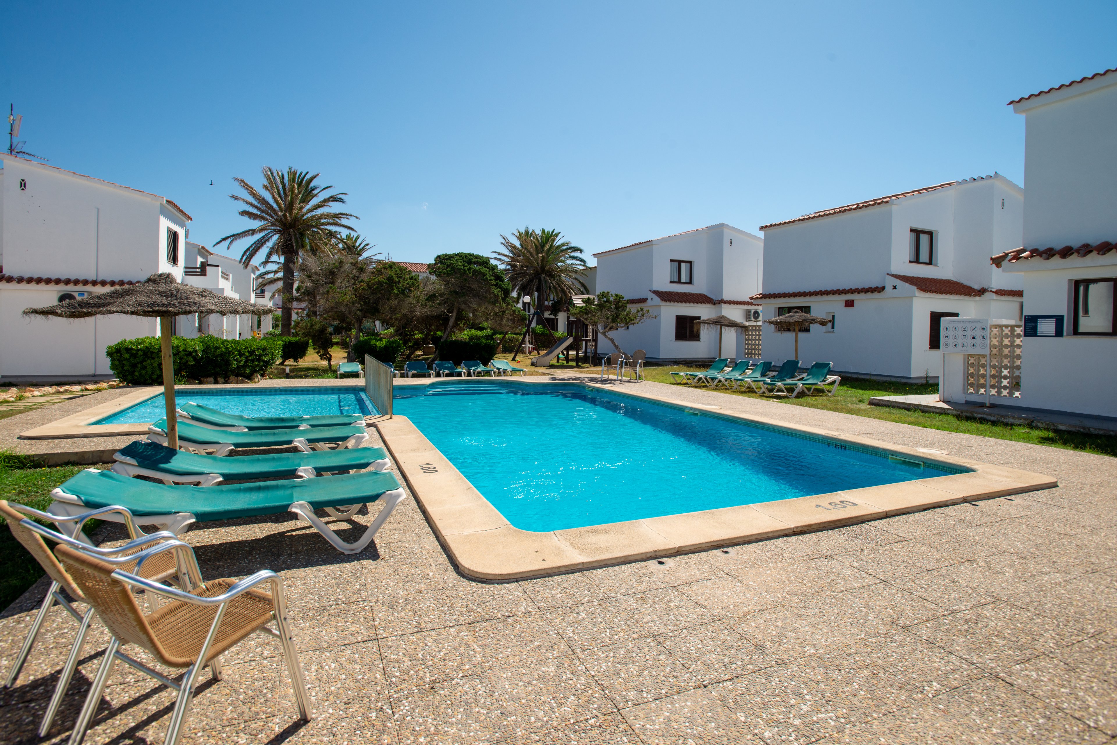 Image of a bright outdoor swimming pool surrounded by sun loungers in a holiday villa complex. White two-story houses with terracotta roofs frame the pool area under a clear blue sky. Palm trees and trimmed hedges add greenery and a Mediterranean feel to the setting. The scene suggests a relaxed vacation atmosphere with ample space for guests to sunbathe and swim.