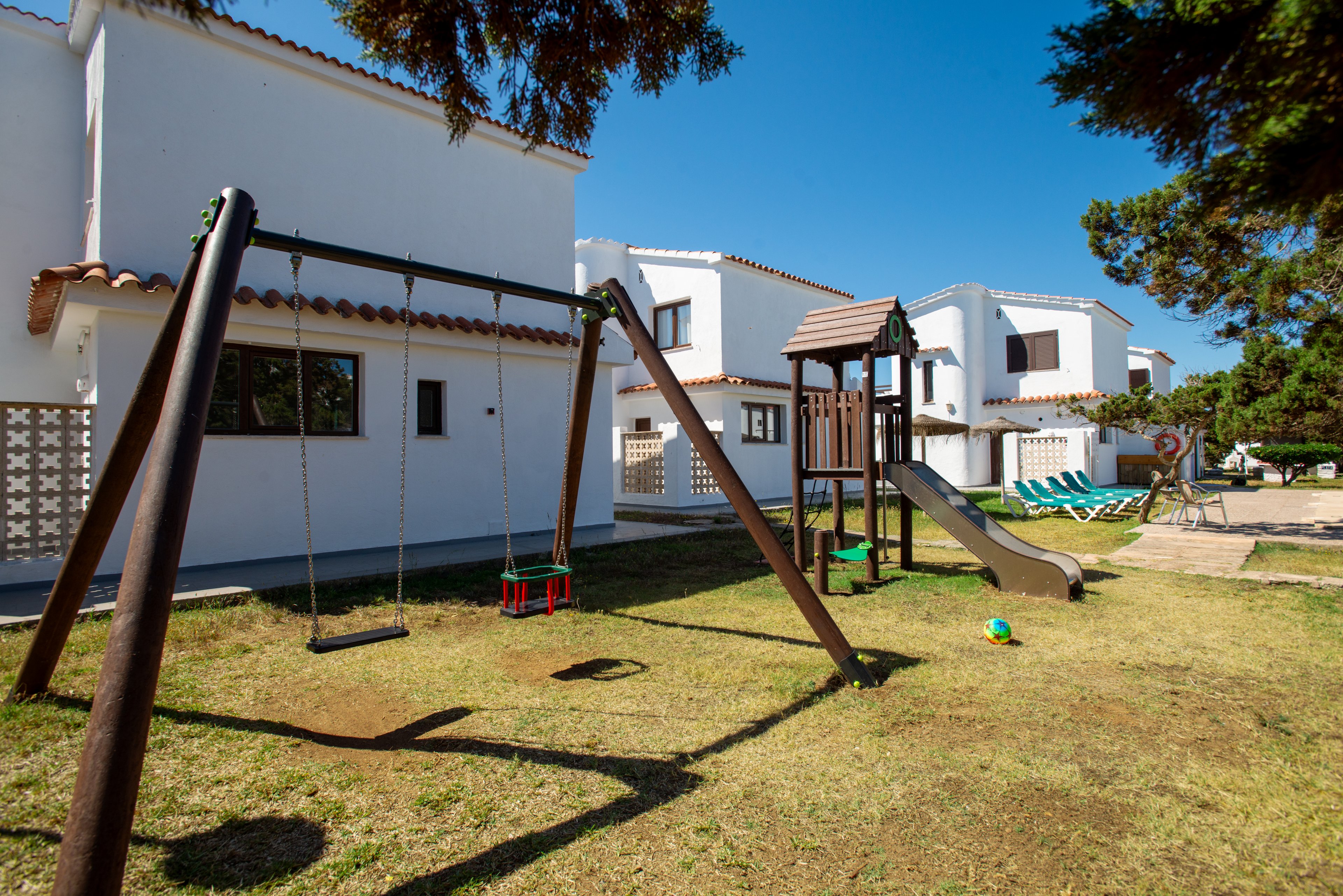 An outdoor playground with swings, a slide, and a small climbing structure stands in front of white residential houses. The scene is set on a grassy yard under clear blue skies, suggesting a warm, sunny day. Several lounge chairs are arranged near the houses, and a colorful ball lies on the grass, adding to the family-friendly atmosphere. Trees frame the area, providing shade and a sense of neighborhood tranquility.