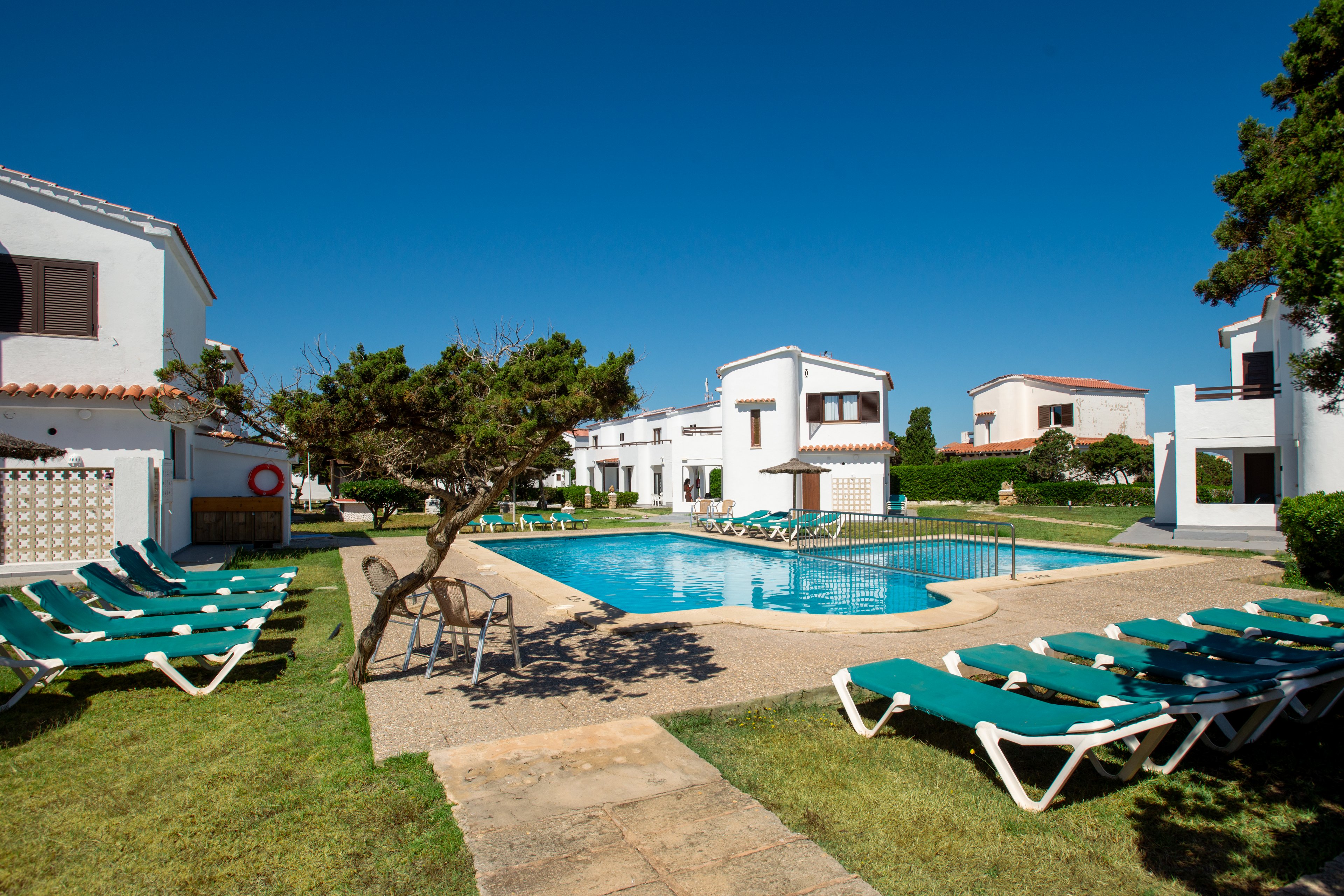 White holiday apartment buildings surround a clear blue outdoor swimming pool under a bright sky. Green sun loungers are neatly arranged on the grass around the pool, suggesting a relaxing vacation setting. A small tree and well-kept lawn add a touch of nature to the scene. The overall atmosphere is calm, sunny, and inviting for guests.