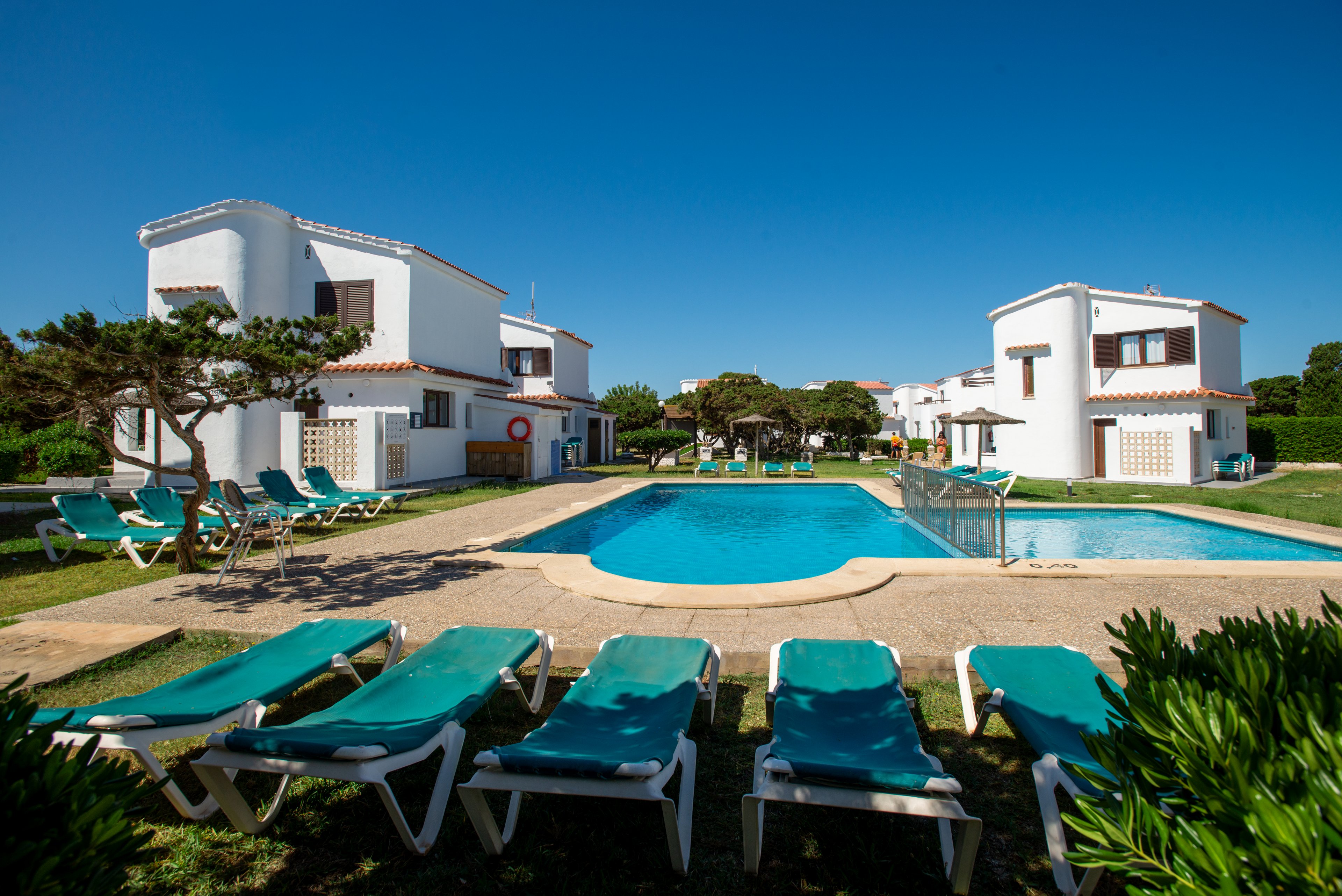 White two-story holiday villas surround a central outdoor swimming pool under a clear blue sky. Green sun loungers line the poolside terrace, suggesting a relaxing vacation setting. Well-kept lawns, shrubs, and a few sculpted trees add a touch of greenery to the scene. The bright daylight and vivid colors create a cheerful, inviting atmosphere for guests.