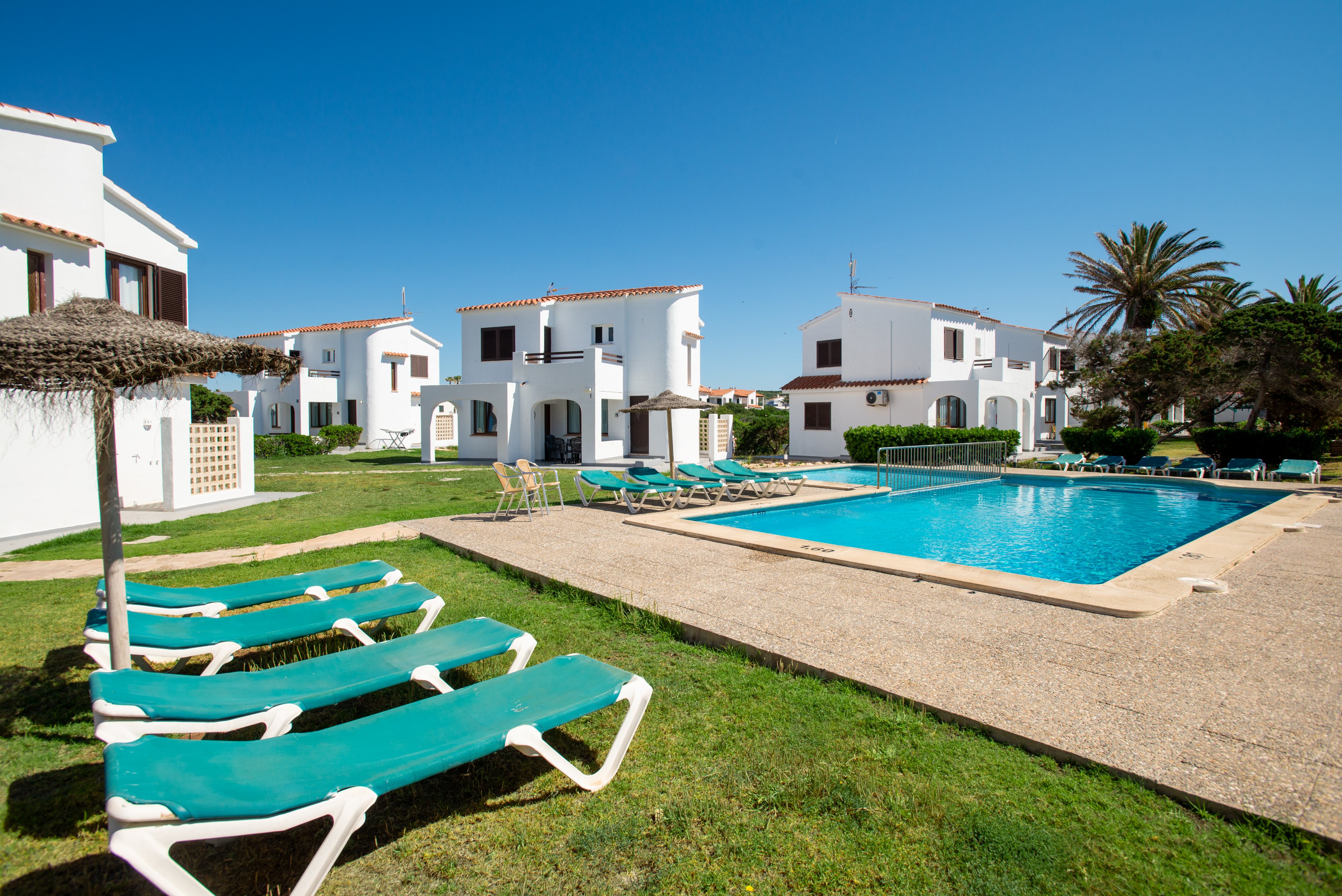 White two-story holiday villas surround a bright blue outdoor swimming pool under a clear sky. Green sun loungers are arranged on the grass and poolside terrace, suggesting a relaxing vacation setting. A large palm tree and neatly maintained lawn add to the Mediterranean resort atmosphere. The scene is empty of people, emphasizing tranquility and leisure.