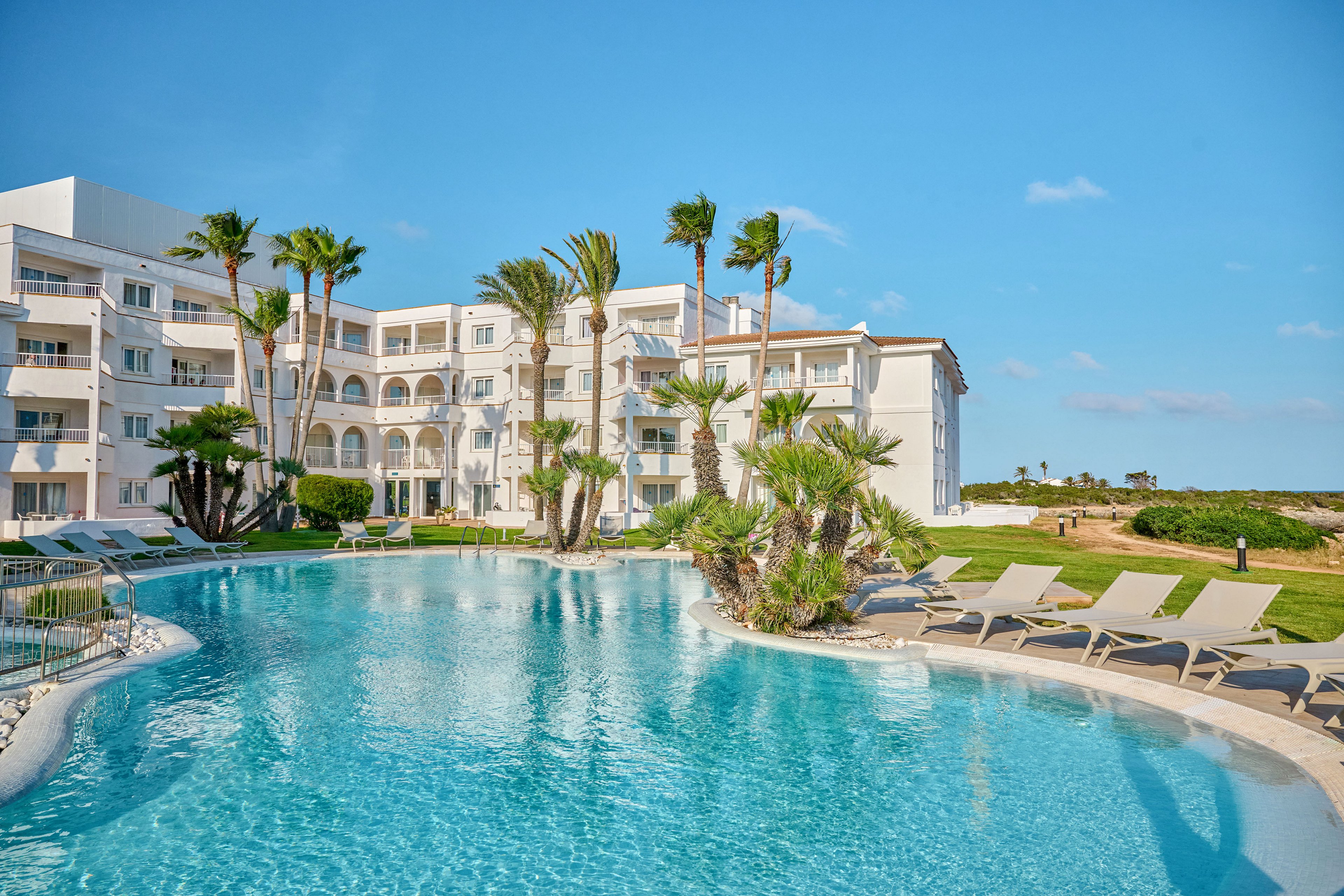 A large outdoor swimming pool sits in front of a white multi-story resort building. Palm trees and manicured lawns surround the pool, creating a tropical vacation atmosphere. Several empty lounge chairs line the poolside under a clear blue sky. The scene suggests a relaxing holiday destination with bright daylight and calm water.